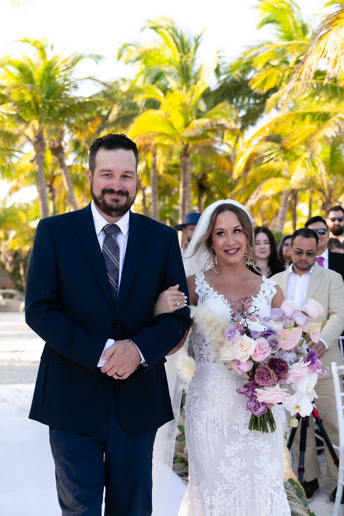 A wedding ceremony takes place on a tropical beach with palm trees and guests gathered under the sun.