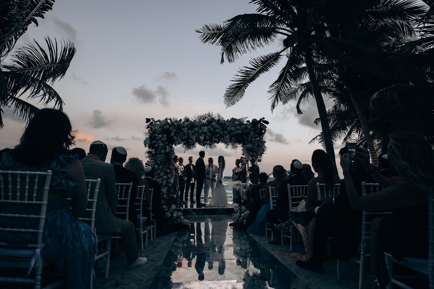 Silhouette of wedding ceremony setup with palm trees against a dramatic evening sky by the ocean.