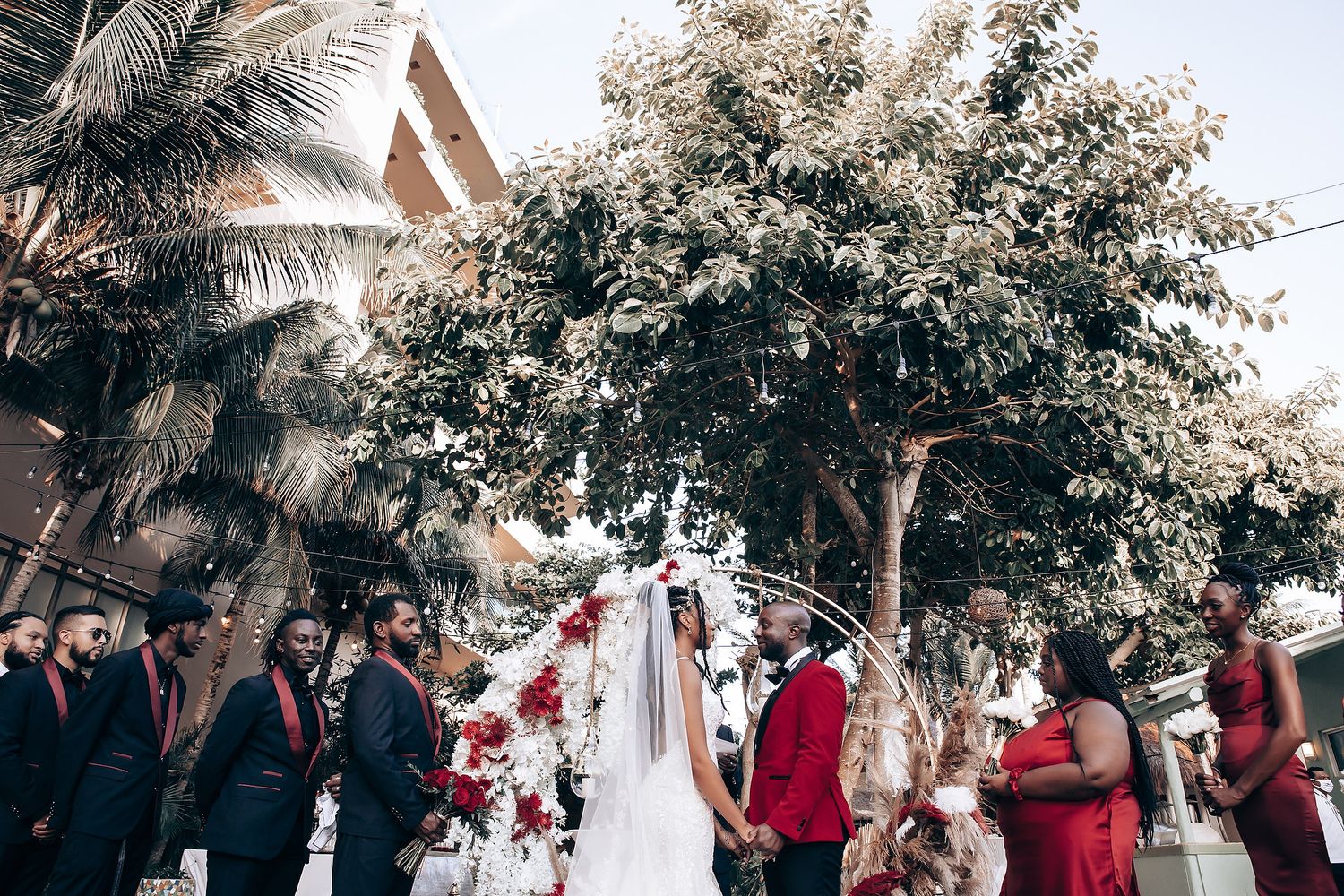 Wedding party gathers under a rustic wooden arch decorated with white flowers and greenery for an outdoor ceremony.