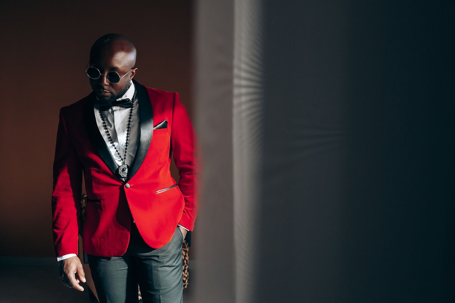 A person in a red blazer and black bowtie stands against a dark wall with dramatic side lighting.