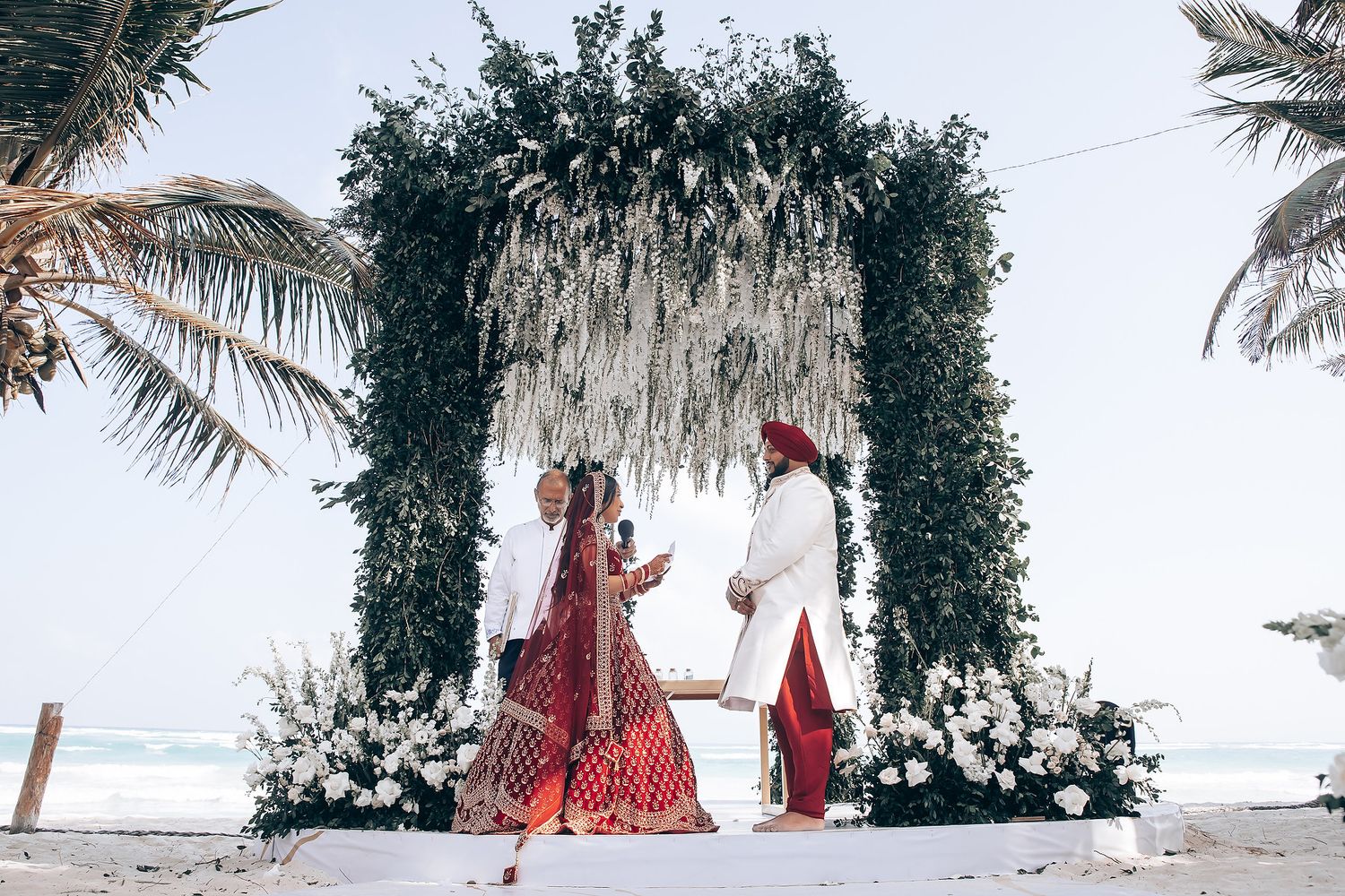 Beach wedding ceremony under floral arch with ocean and palm trees in background.