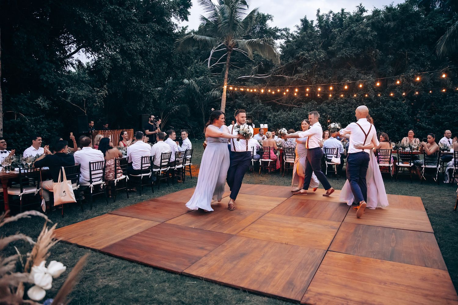 Outdoor wedding reception setup with string lights and wooden dance floor surrounded by trees.