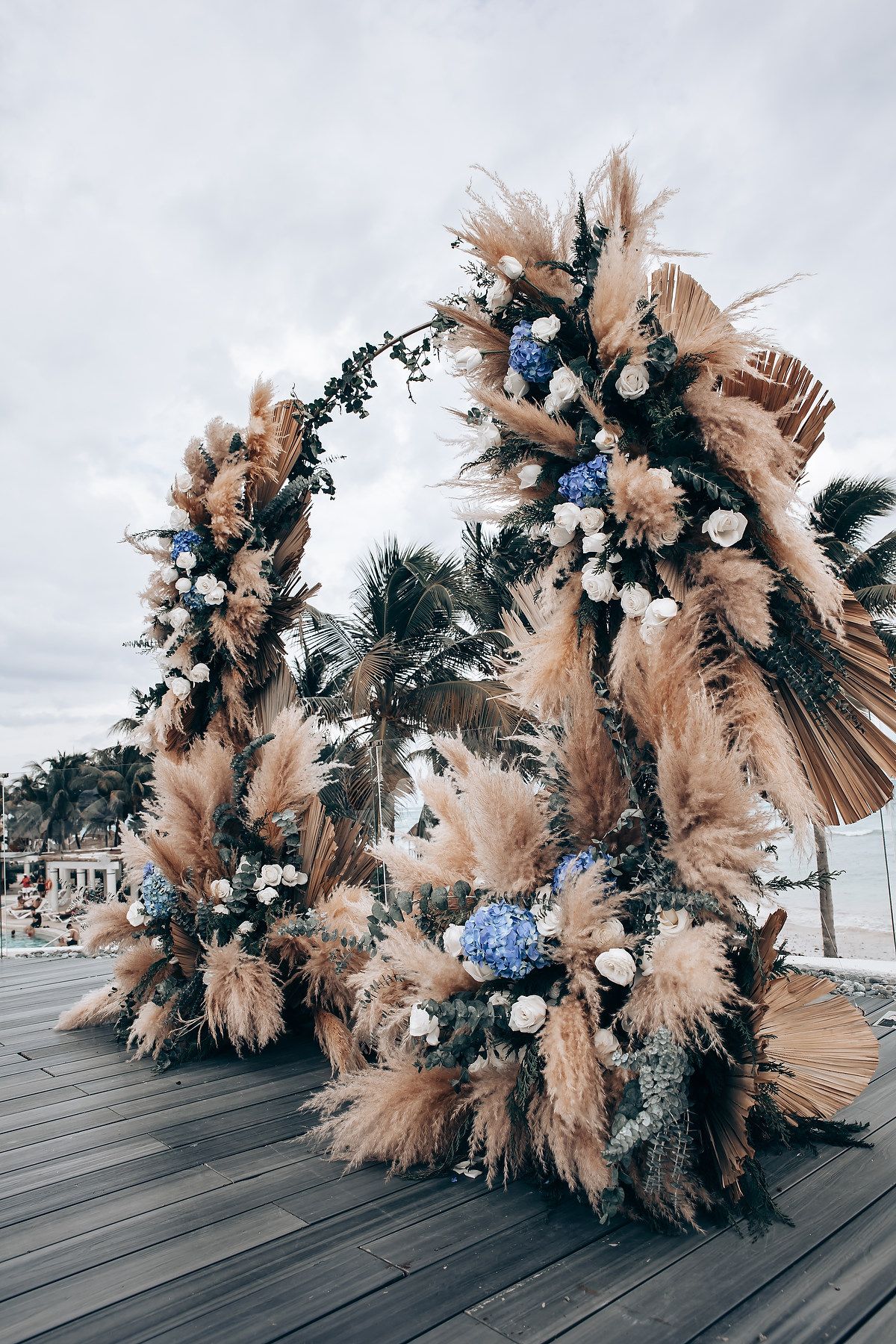 Stunning boho wedding arch with dried pampas grass and blue florals on wooden deck.