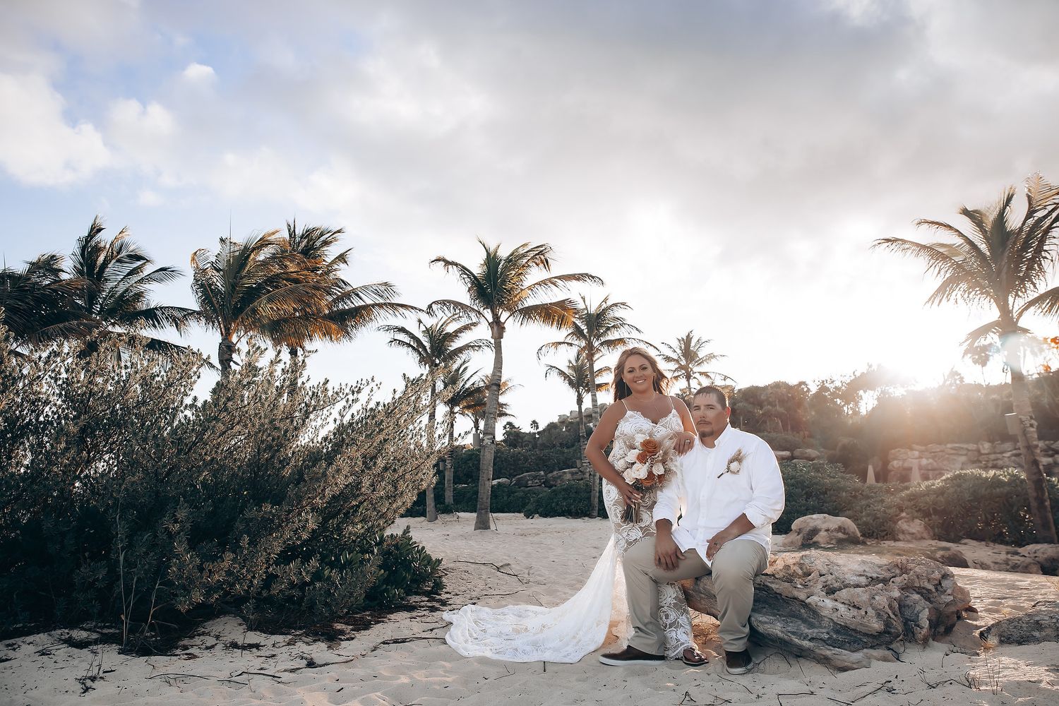 A couple sits on rocky beach at sunset with palm trees swaying in the wind in Hawaii.