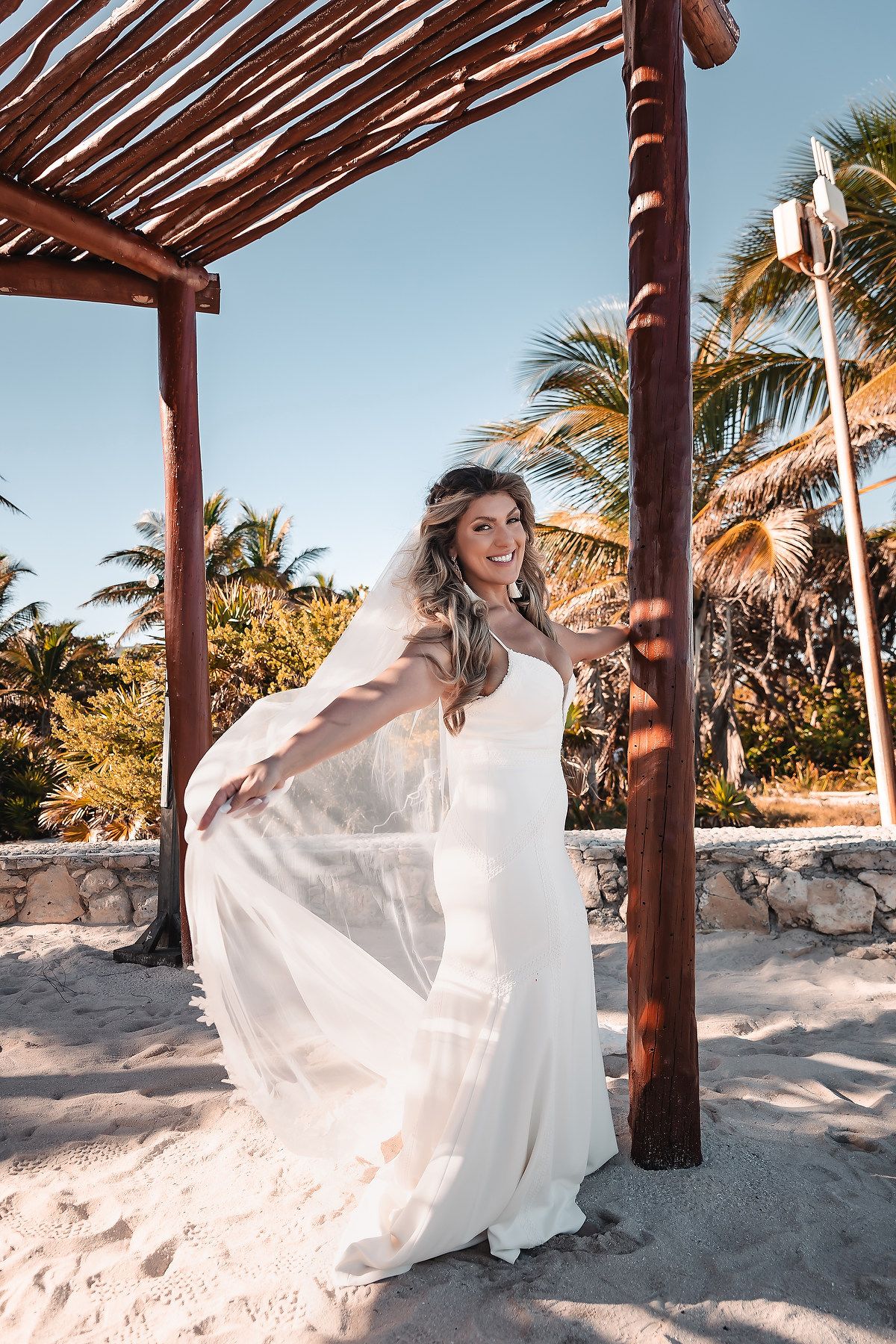 A flowing white wedding dress billows in the breeze under a wooden pergola on a tropical beach with palm trees.