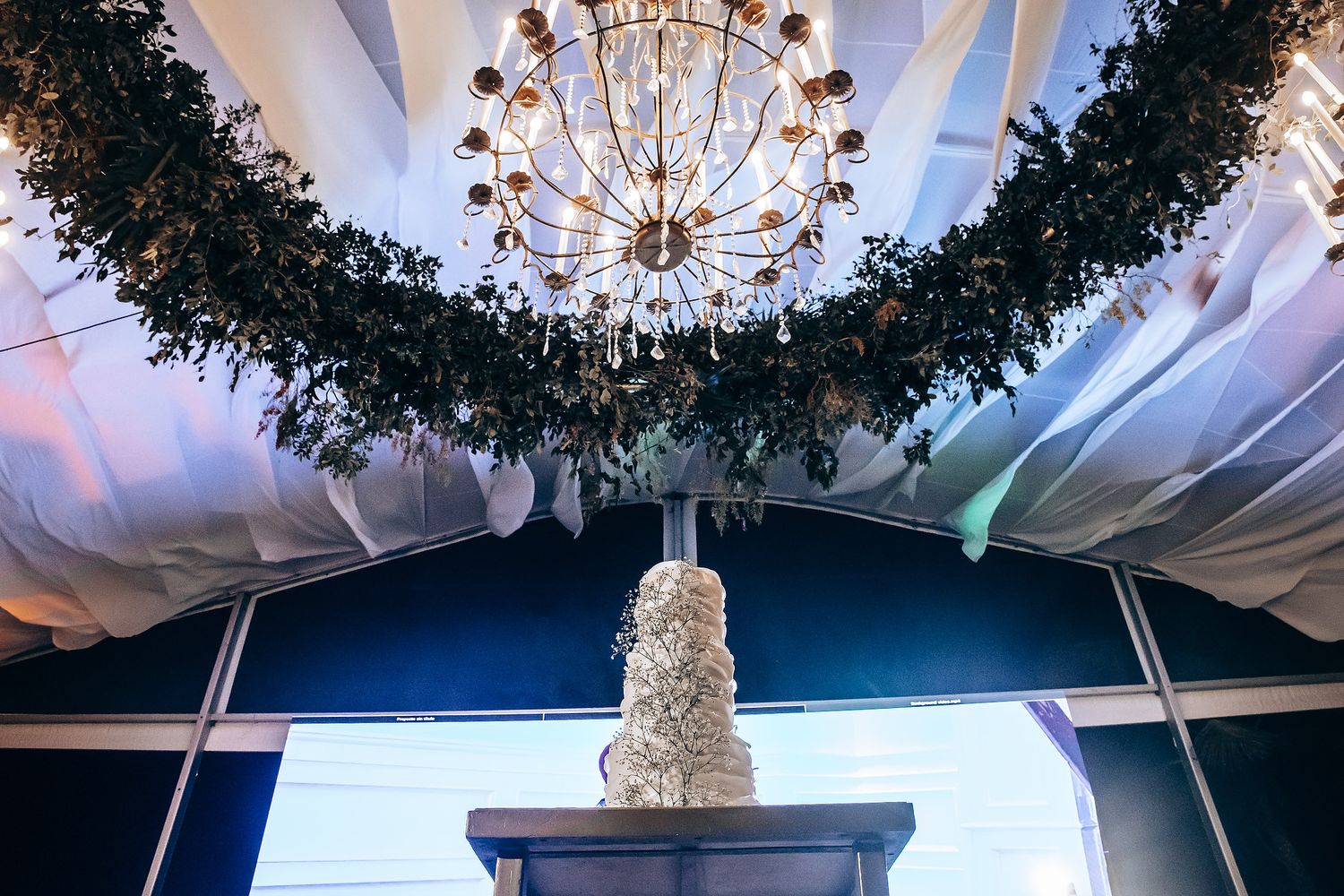 Elegant wedding cake displayed under a crystal chandelier with greenery garland decorating the tent ceiling.