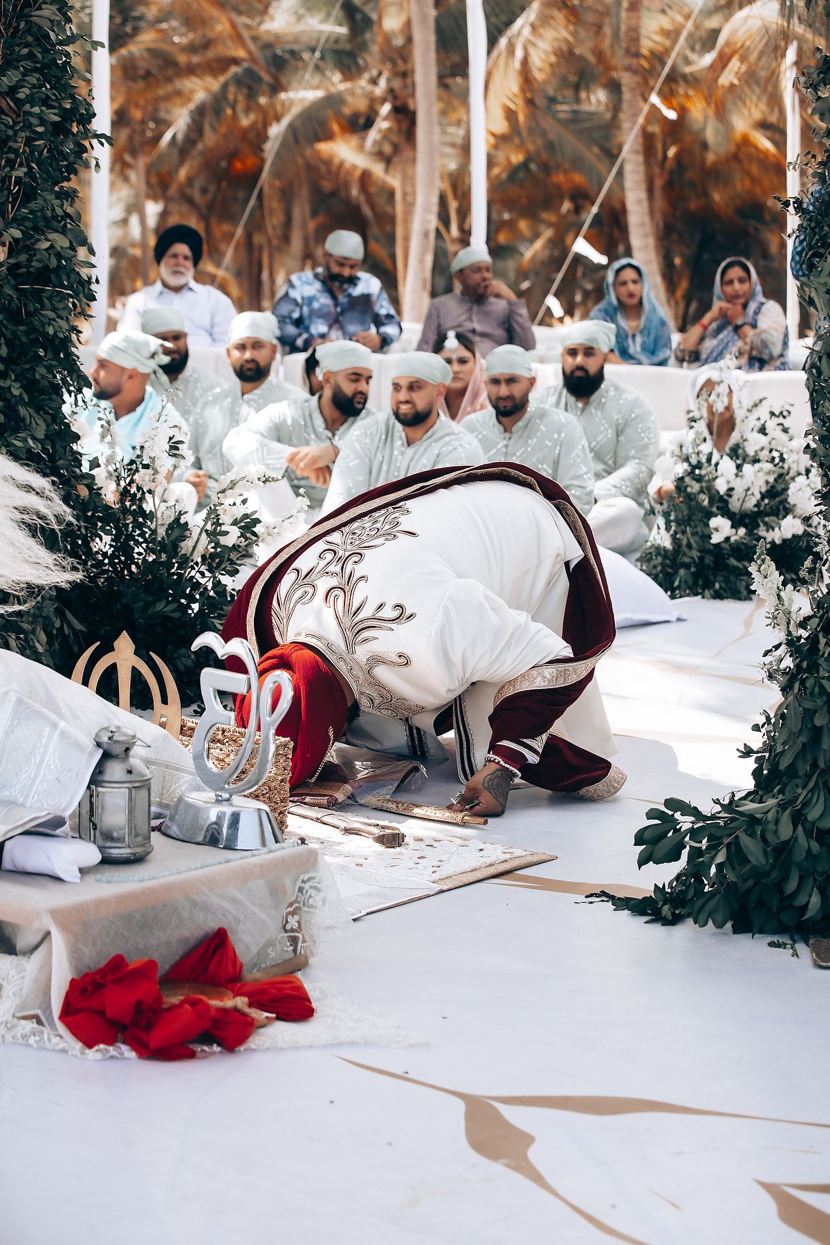 Wedding guests in traditional white attire seated on floor during cultural ceremony with burgundy accents.