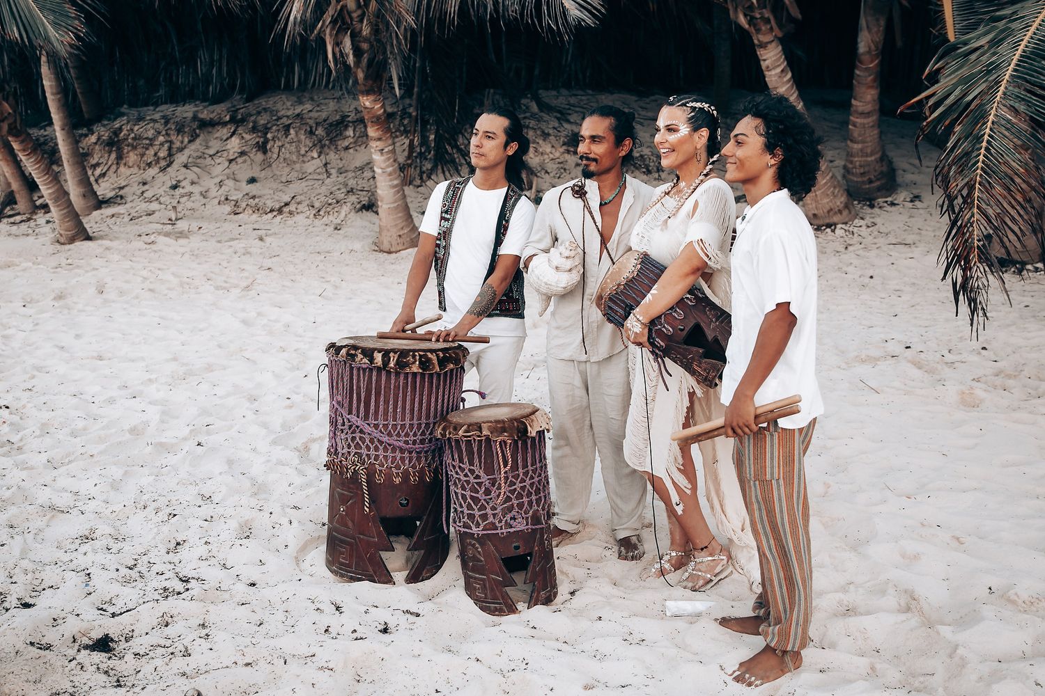 Musicians with drums perform on a tropical beach wearing white and neutral colored attire.