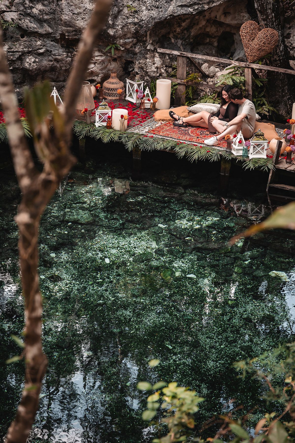 Natural cenote pool reflecting lush greenery with romantic picnic setup along rocky ledge.