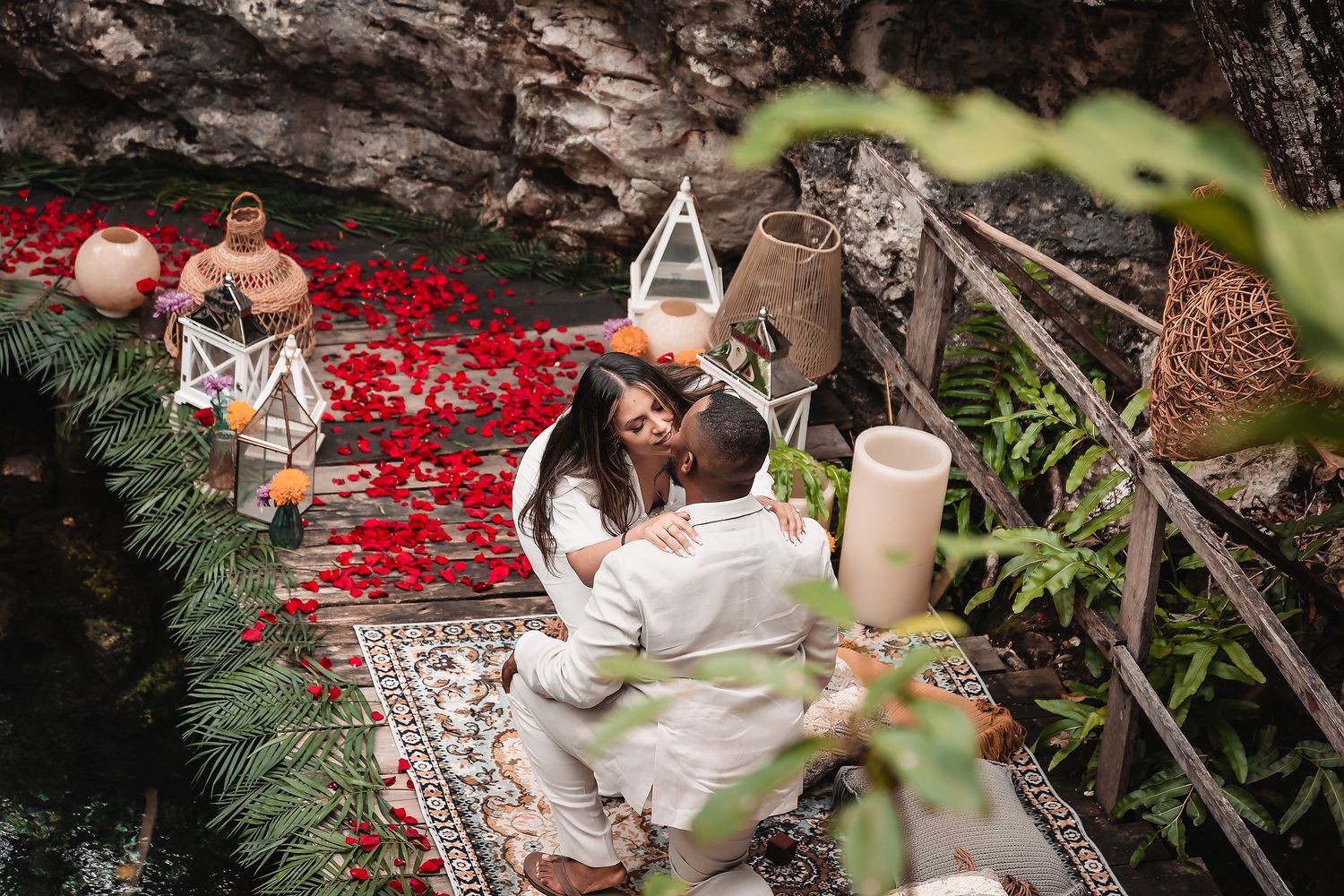 Romantic picnic setup with candles, flowers and decor items on a red blanket in a natural outdoor setting.