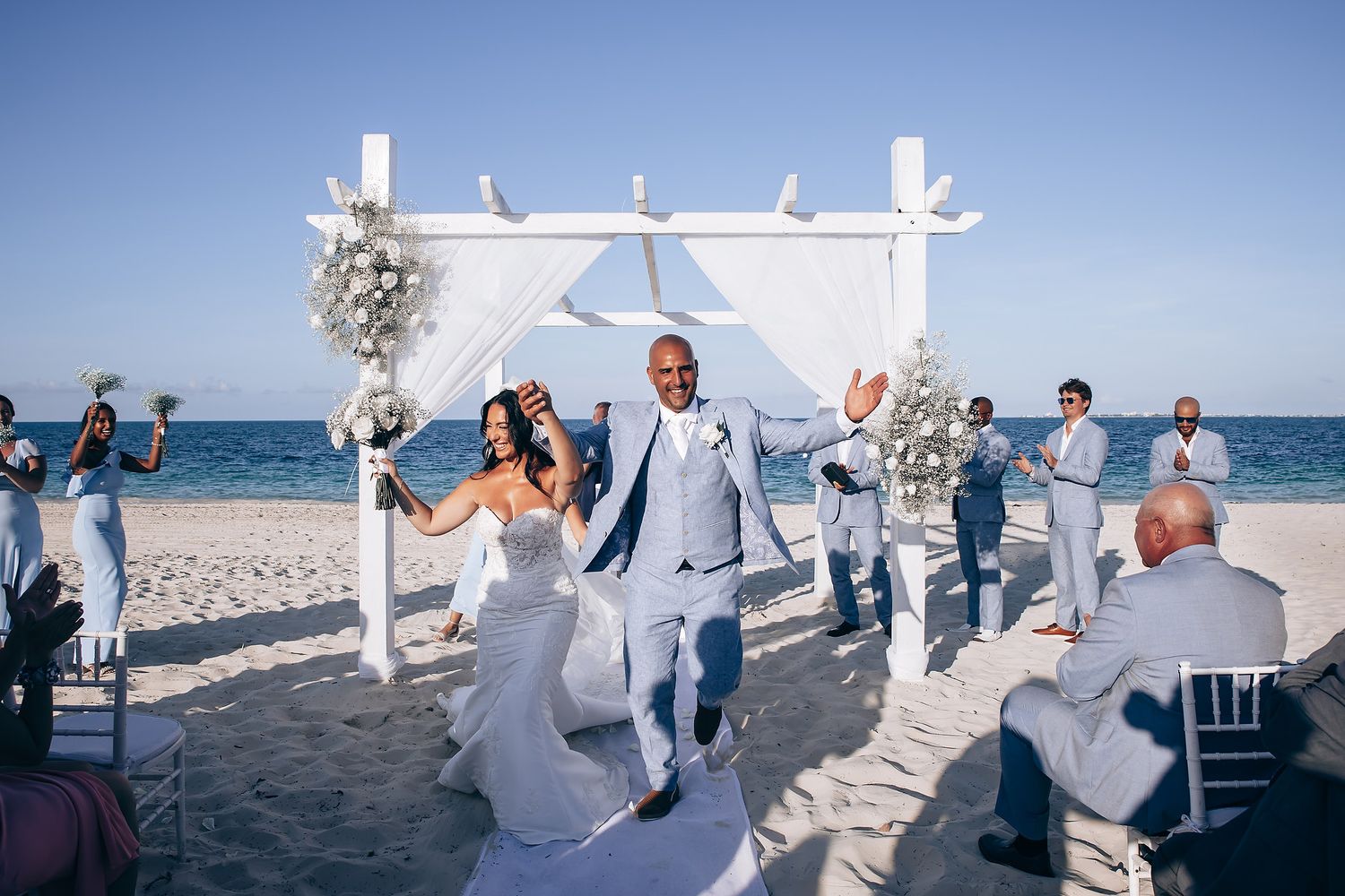 Wedding ceremony site on sandy beach with white fabric draping from wooden structure as guests gather.