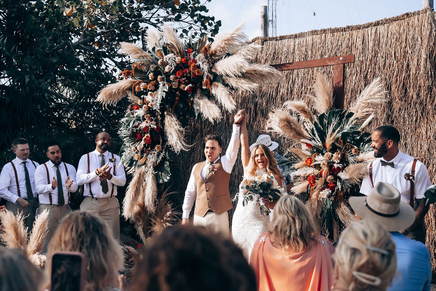 Wedding guests gather outdoors under natural boho decor with dried pampas grass arrangements.