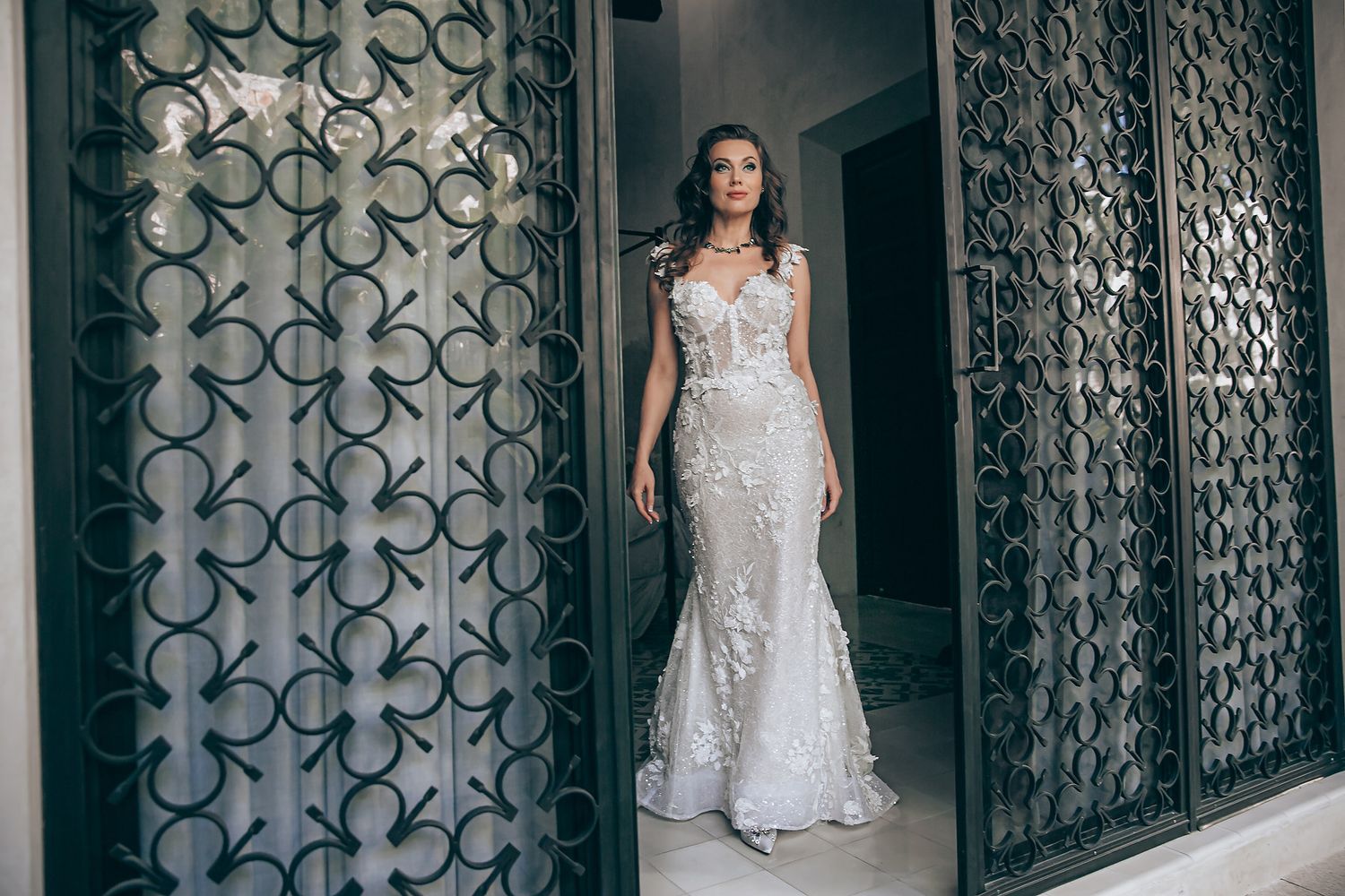 A bride in a flowing white lace gown poses in front of an ornate iron gate with geometric patterns.
