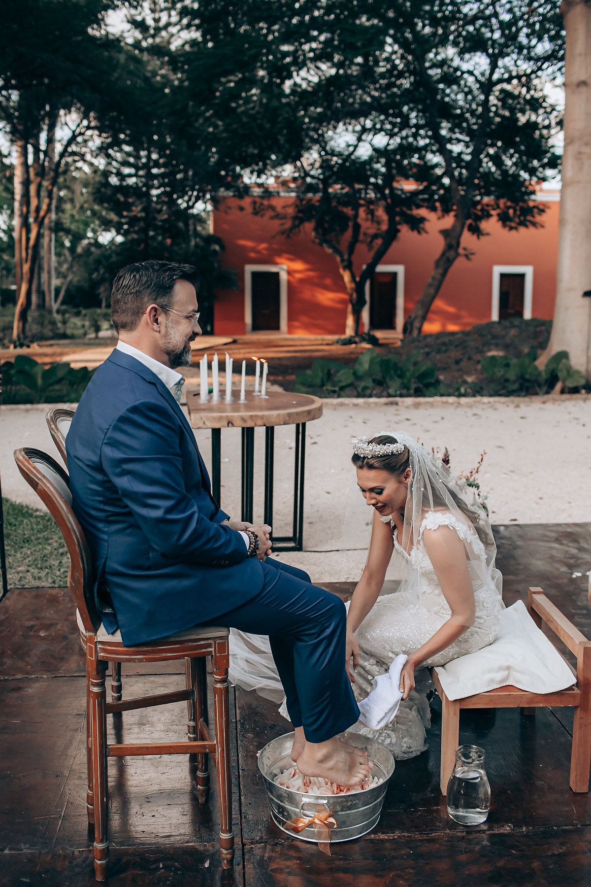 Couple plays traditional wedding shoe game at outdoor venue with rustic wooden chairs.
