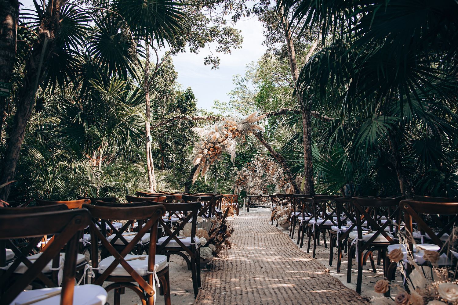 Rustic wooden benches line a palm-tree covered outdoor ceremony venue with natural tropical landscaping.