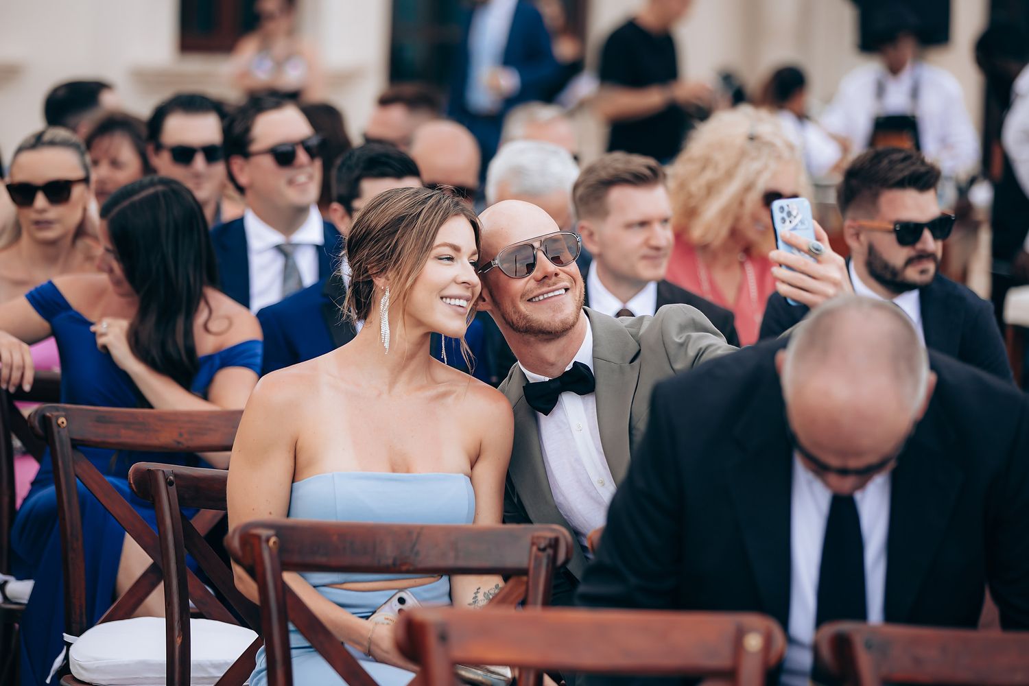 Wedding guests sit at outdoor wooden tables during a daytime reception celebration.