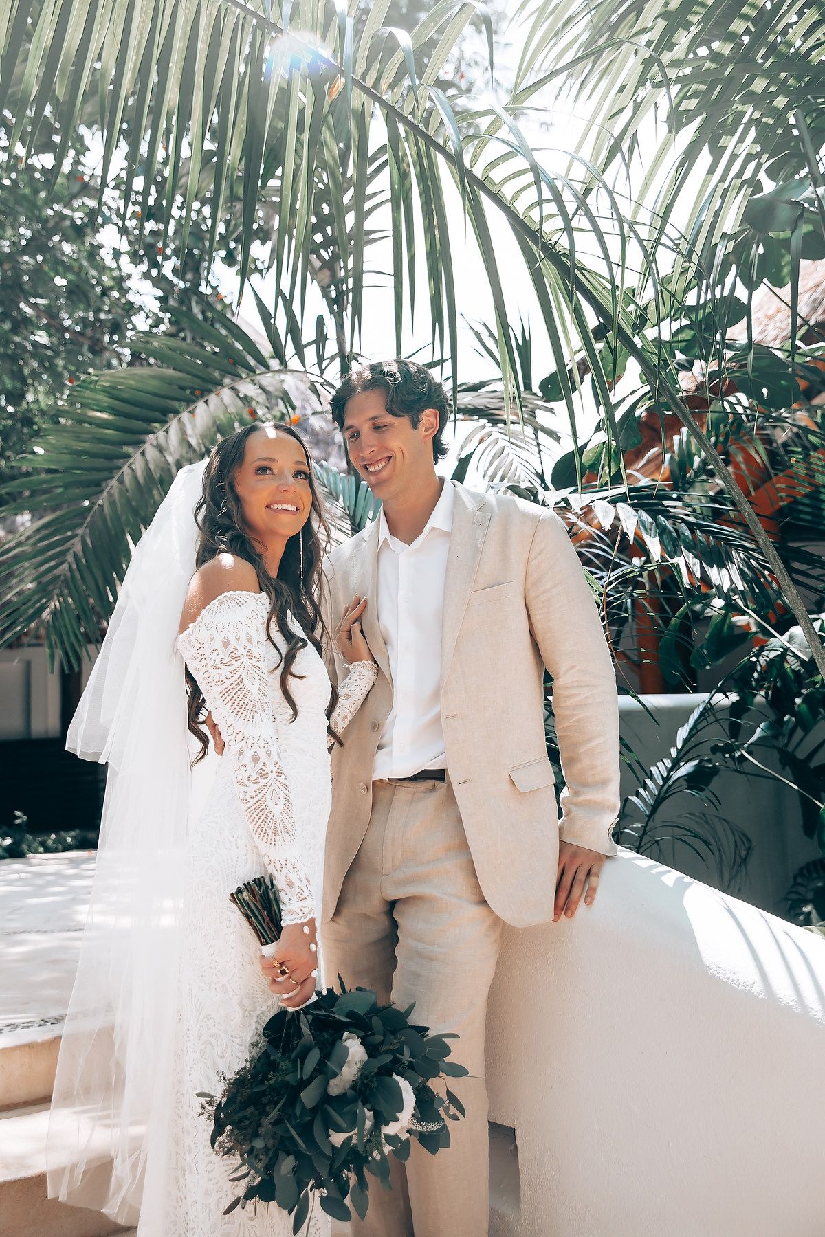 Couple in matching white and beige outfits pose among tropical foliage at their wedding.