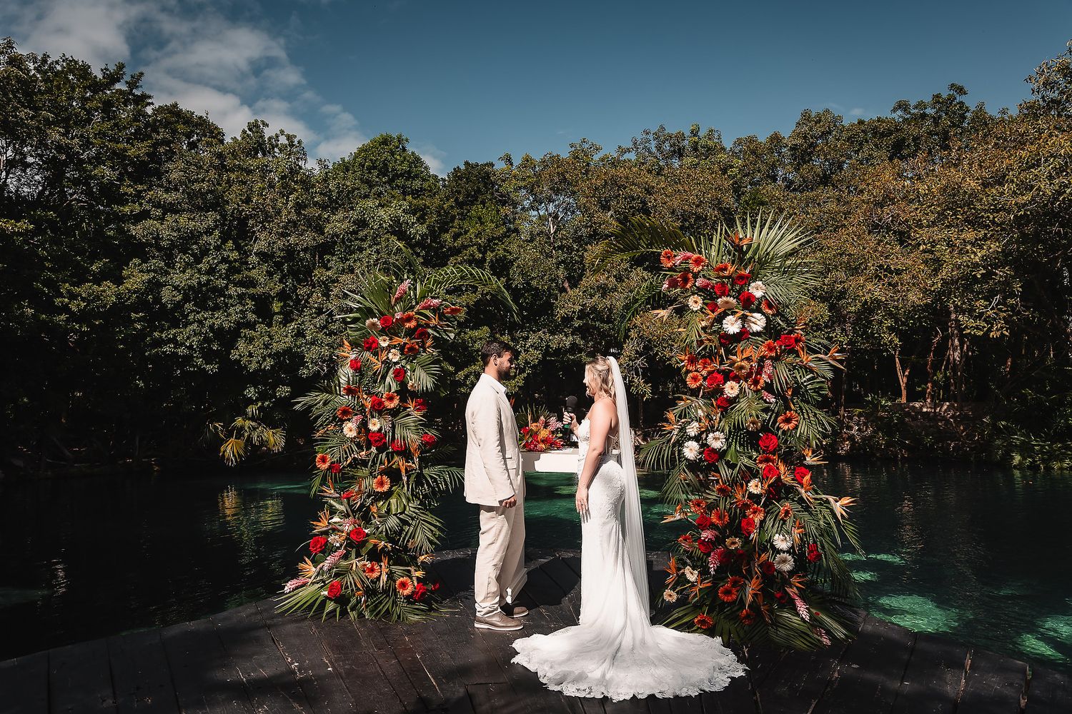 Wedding ceremony setup with stunning red floral arches reflecting in turquoise waters.