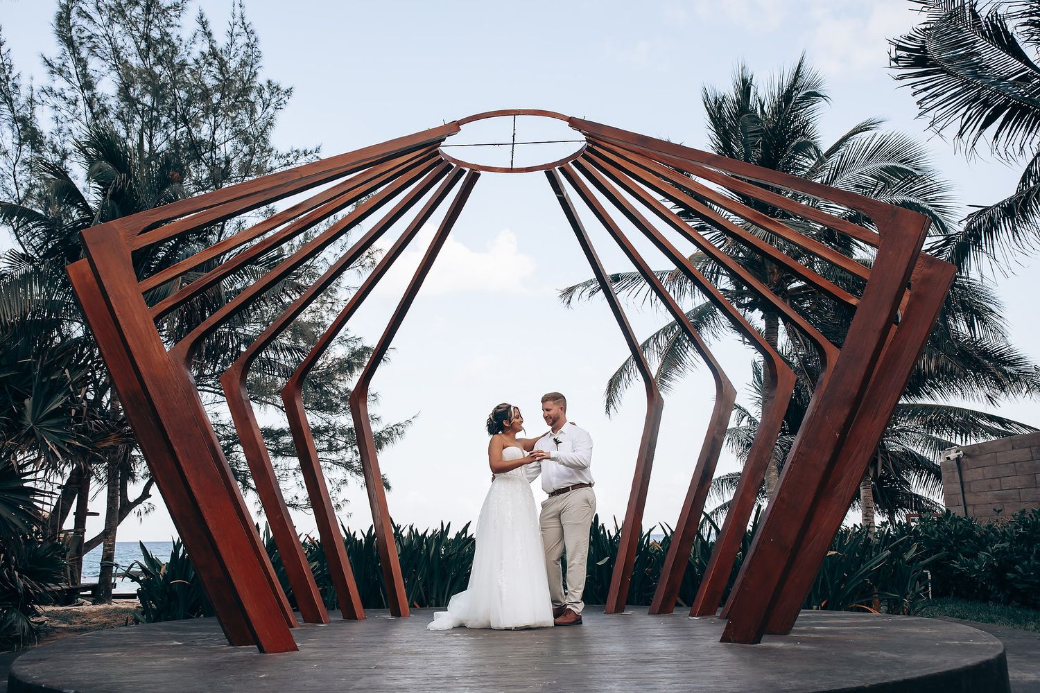 A wedding couple stands beneath a modern wooden gazebo structure with radiating beams against palm trees.