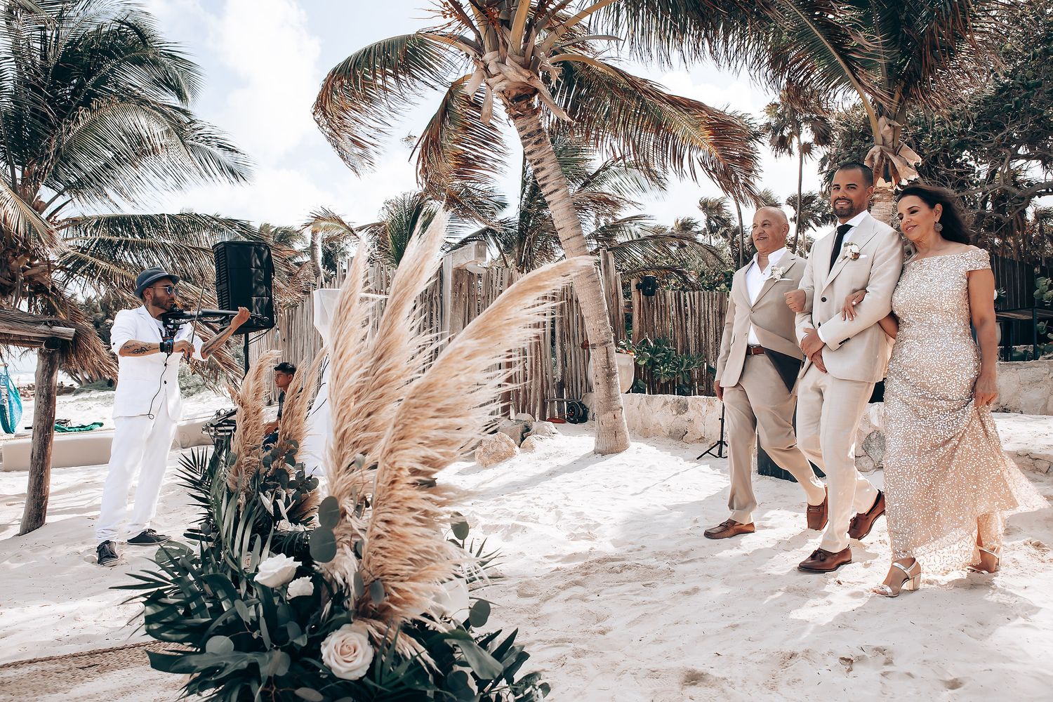 Tropical beach wedding celebration with palm trees and pampas grass decorations on sandy shore.