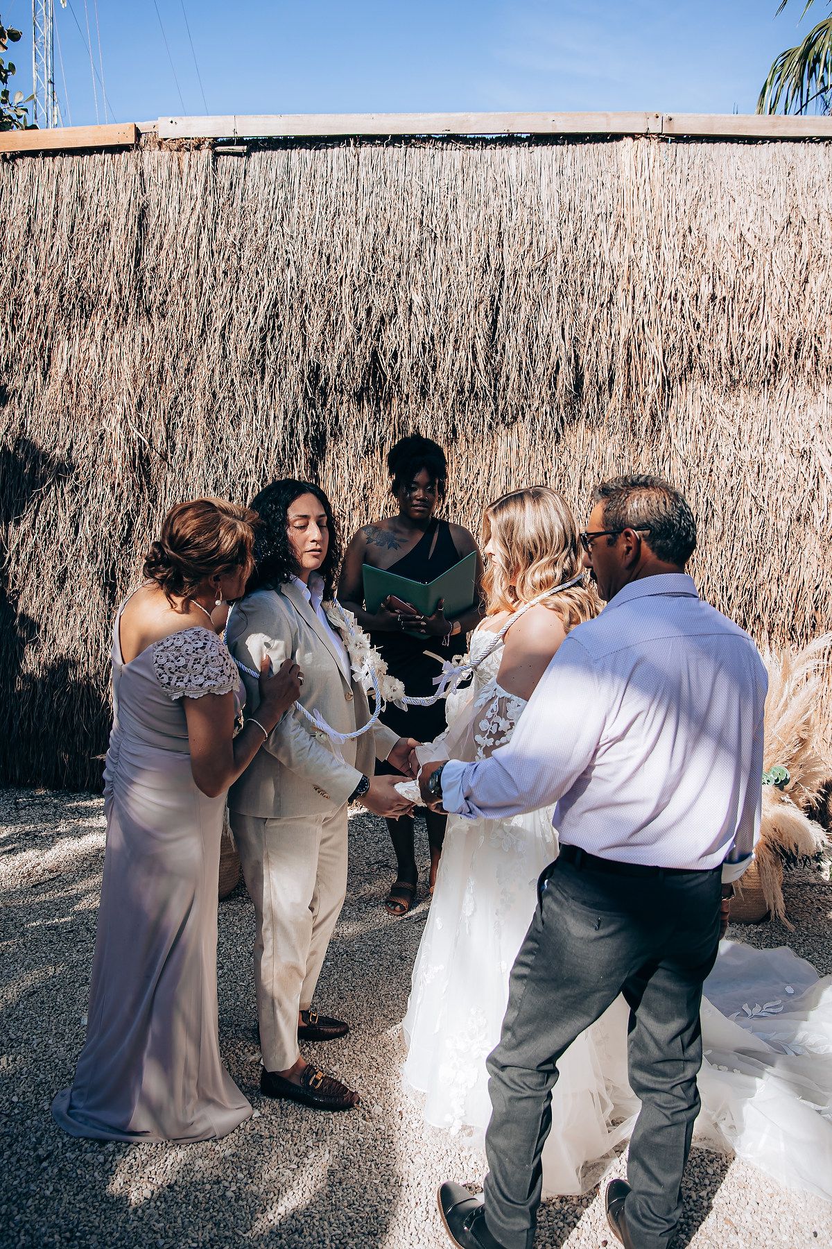 A group dances together at an outdoor celebration with a rustic backdrop of dried reeds or grasses.
