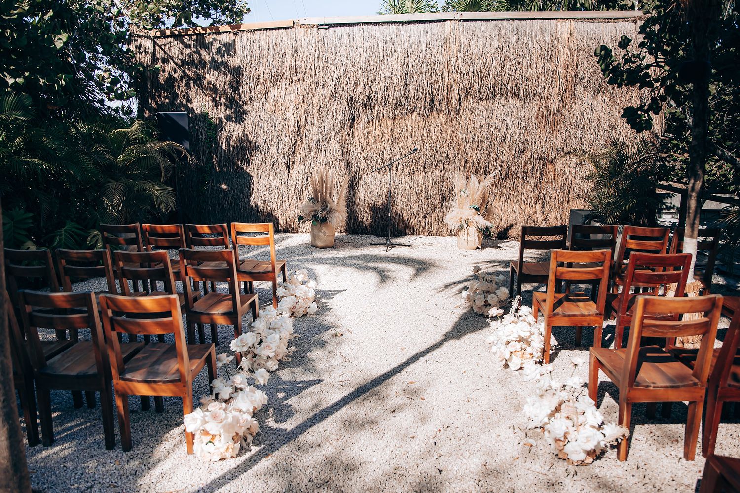 Rustic outdoor dining area with wooden tables and chairs under a thatched roof pergola in a tropical setting.