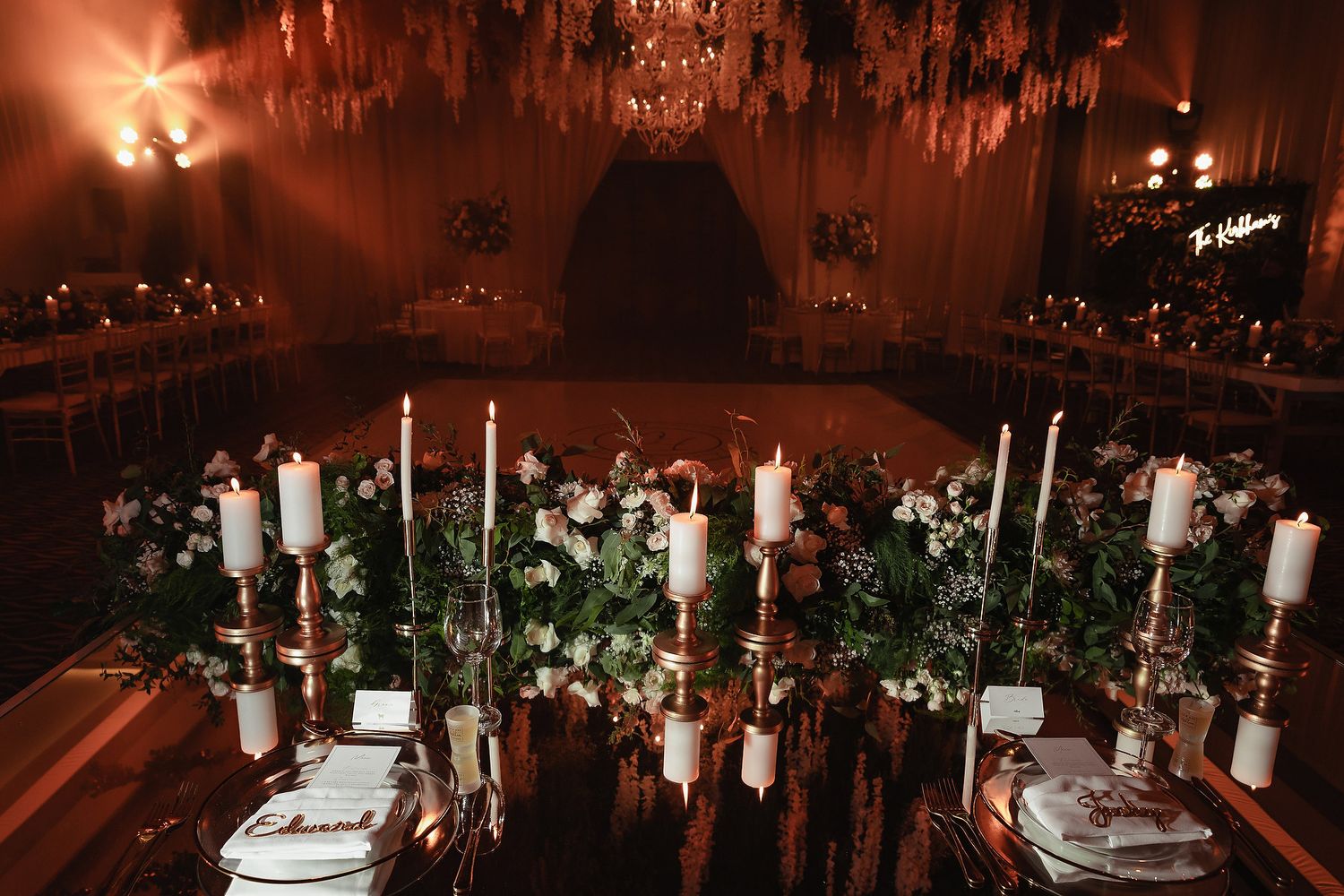 Elegant wedding reception table decorated with white candles and greenery under warm chandelier lighting in dark room.