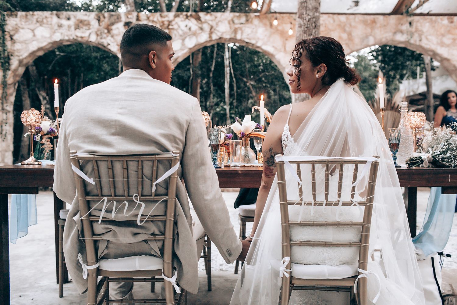 Wedding guests seated in white chairs at an outdoor reception area with natural decor elements.