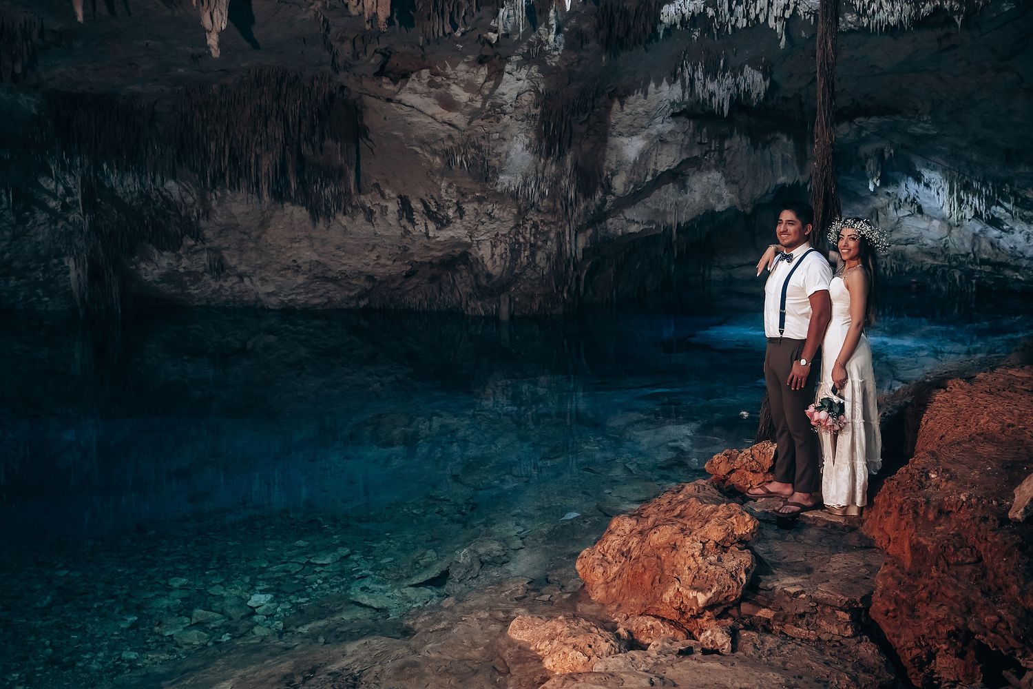 A wedding couple stands near stunning turquoise waters inside a dramatic cave formation.