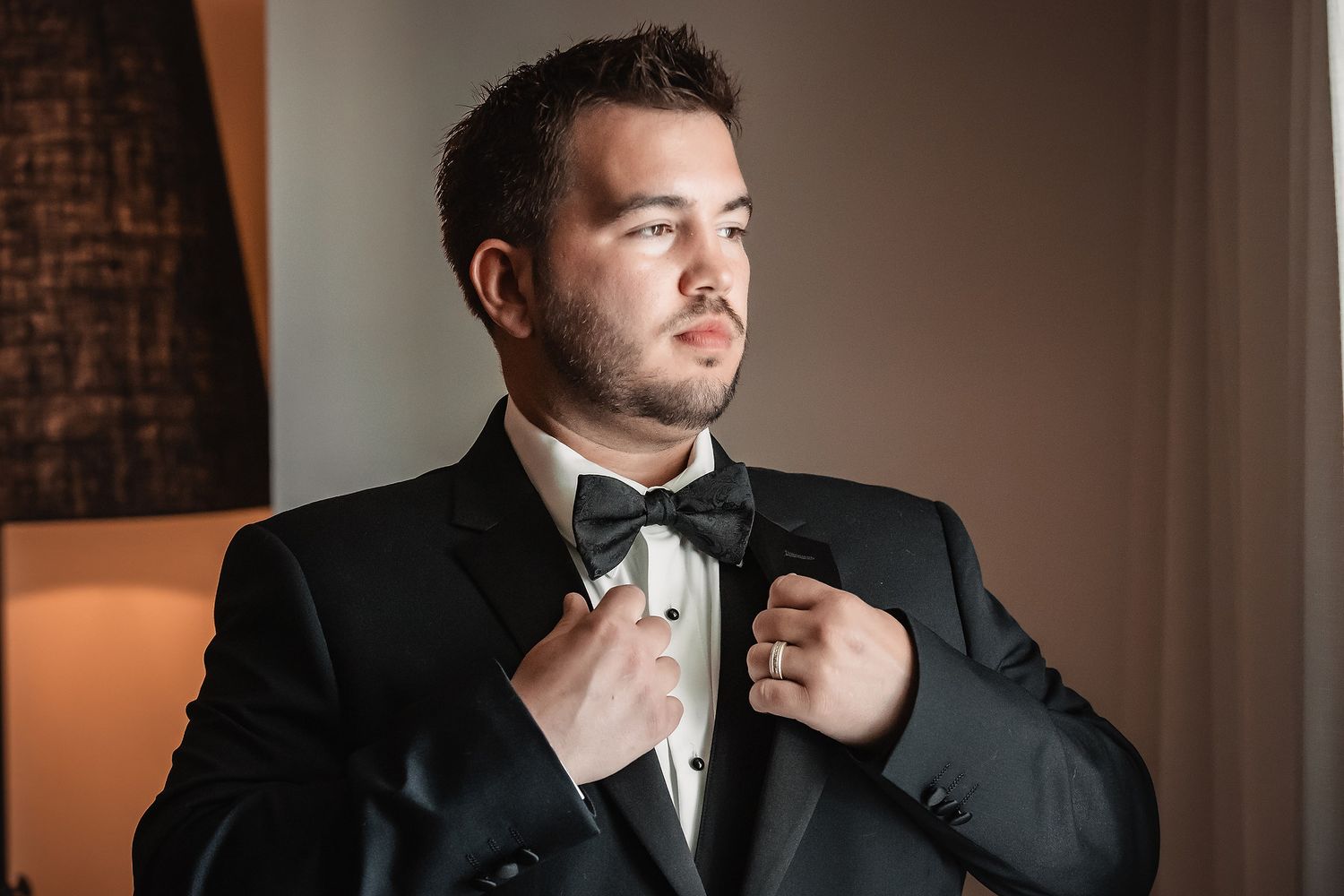 A person in a black tuxedo and bow tie adjusts their collar while standing in a dimly lit room.