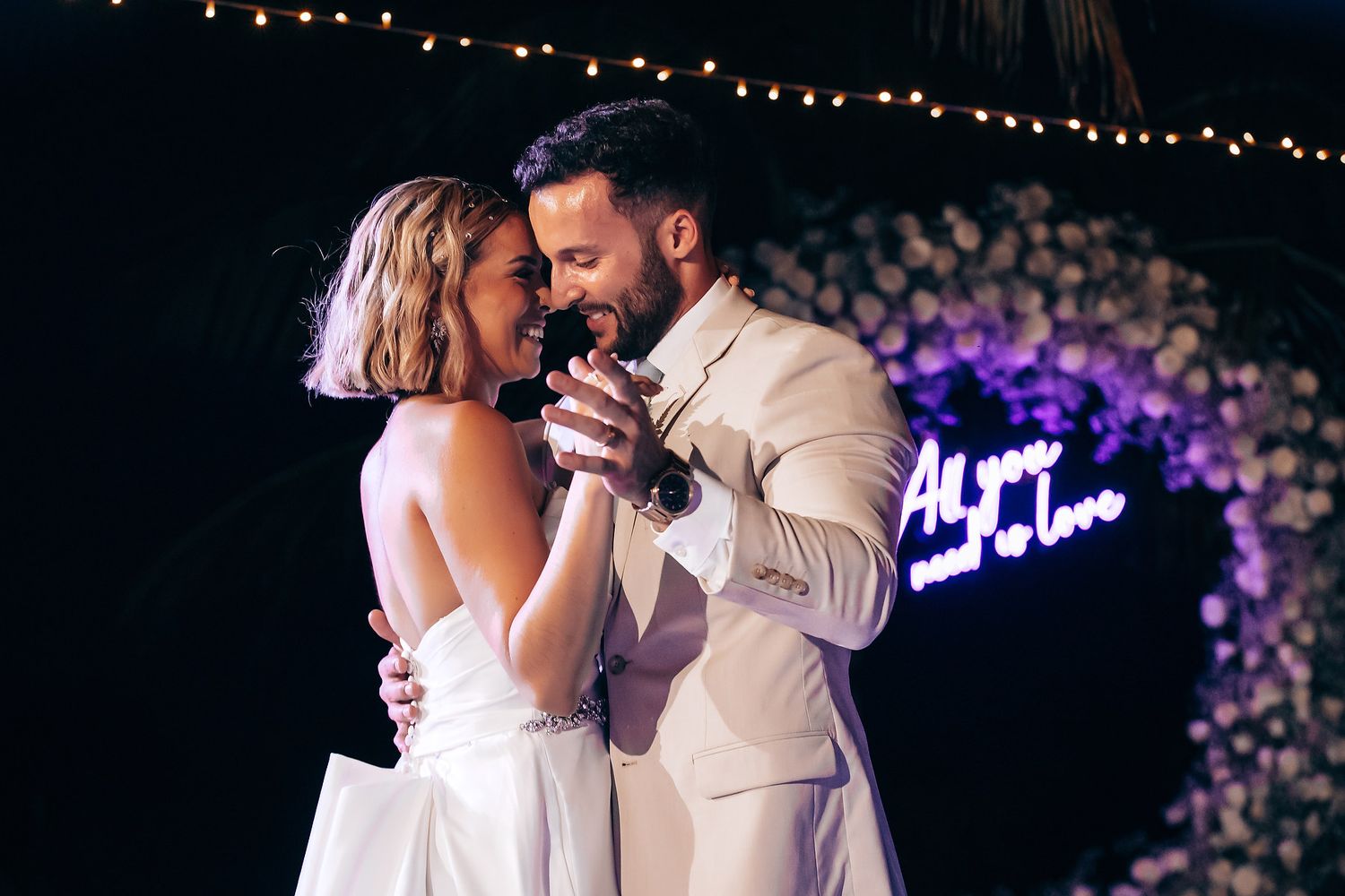 Couple dancing under purple floral arch and neon lights at evening reception.