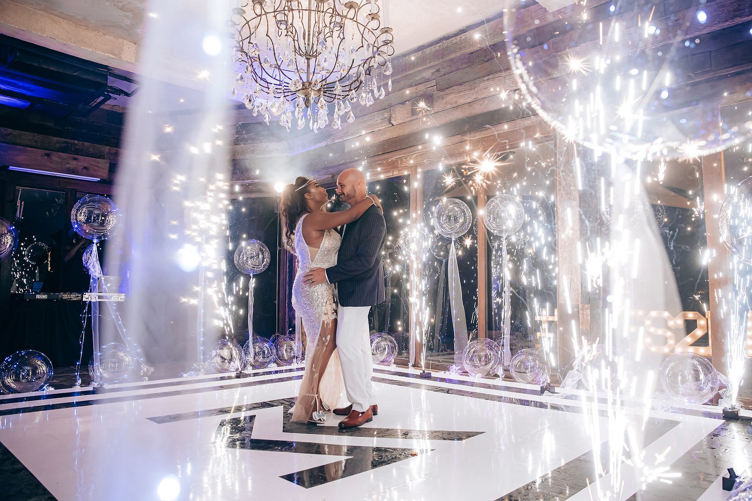 A couple shares their first dance on a reflective dance floor surrounded by sparklers and elegant crystal chandeliers.