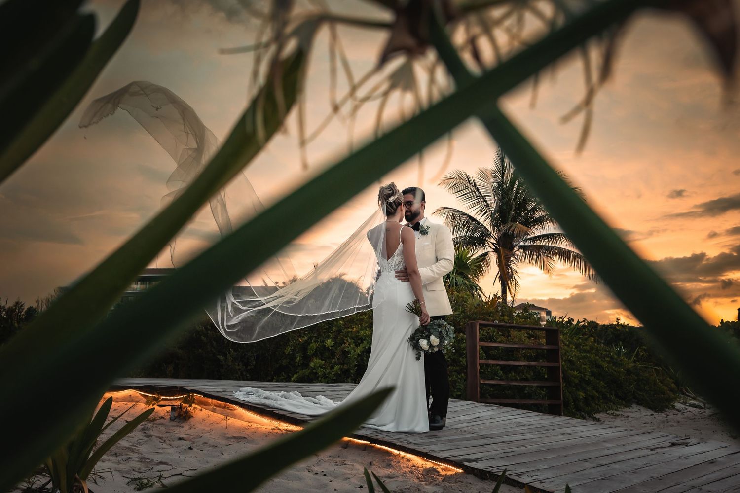 A romantic sunset wedding photo captured through palm fronds on a tropical beach with dramatic lighting.