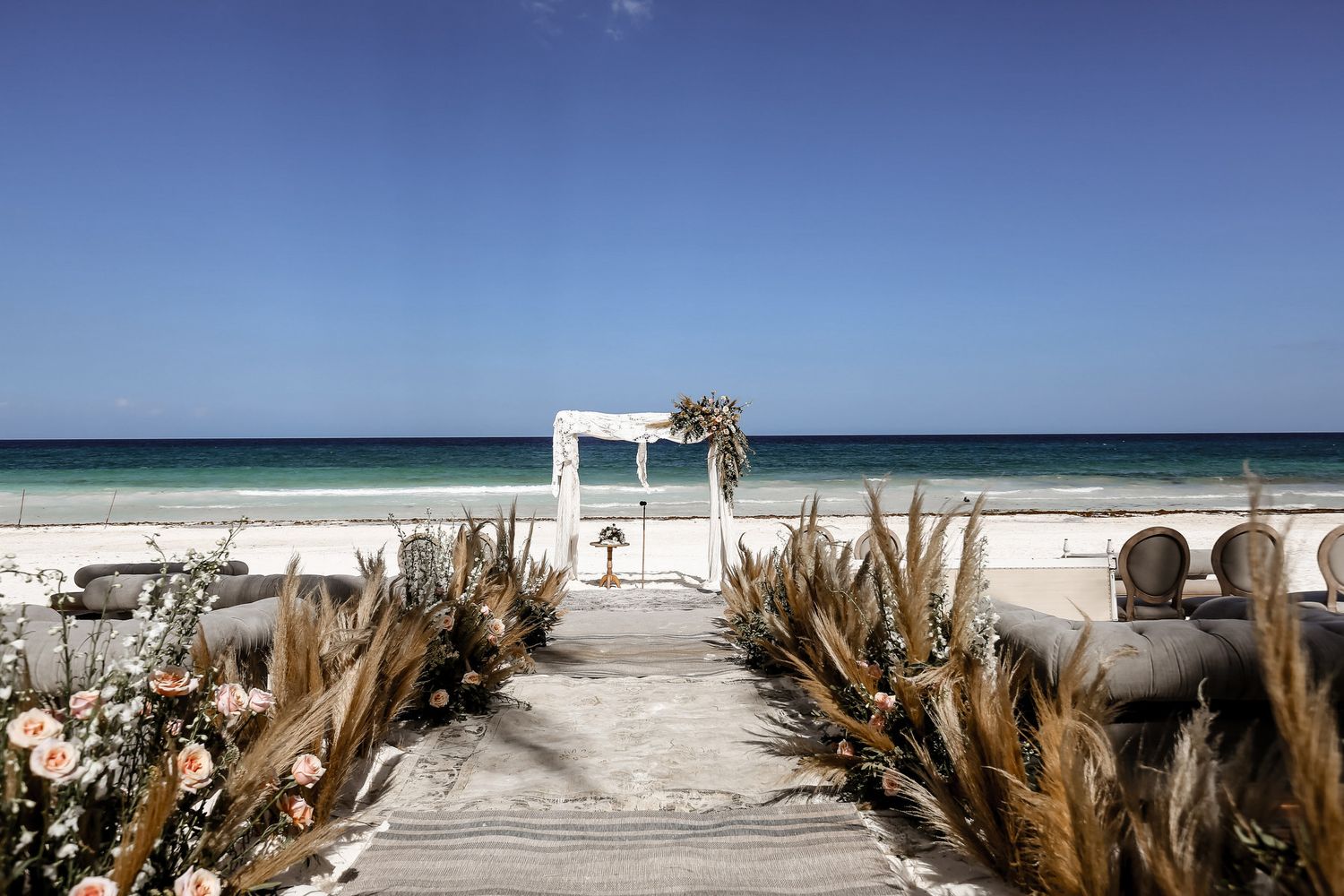 Tropical beach wedding venue with white sand, blue ocean, and pampas grass decorations.