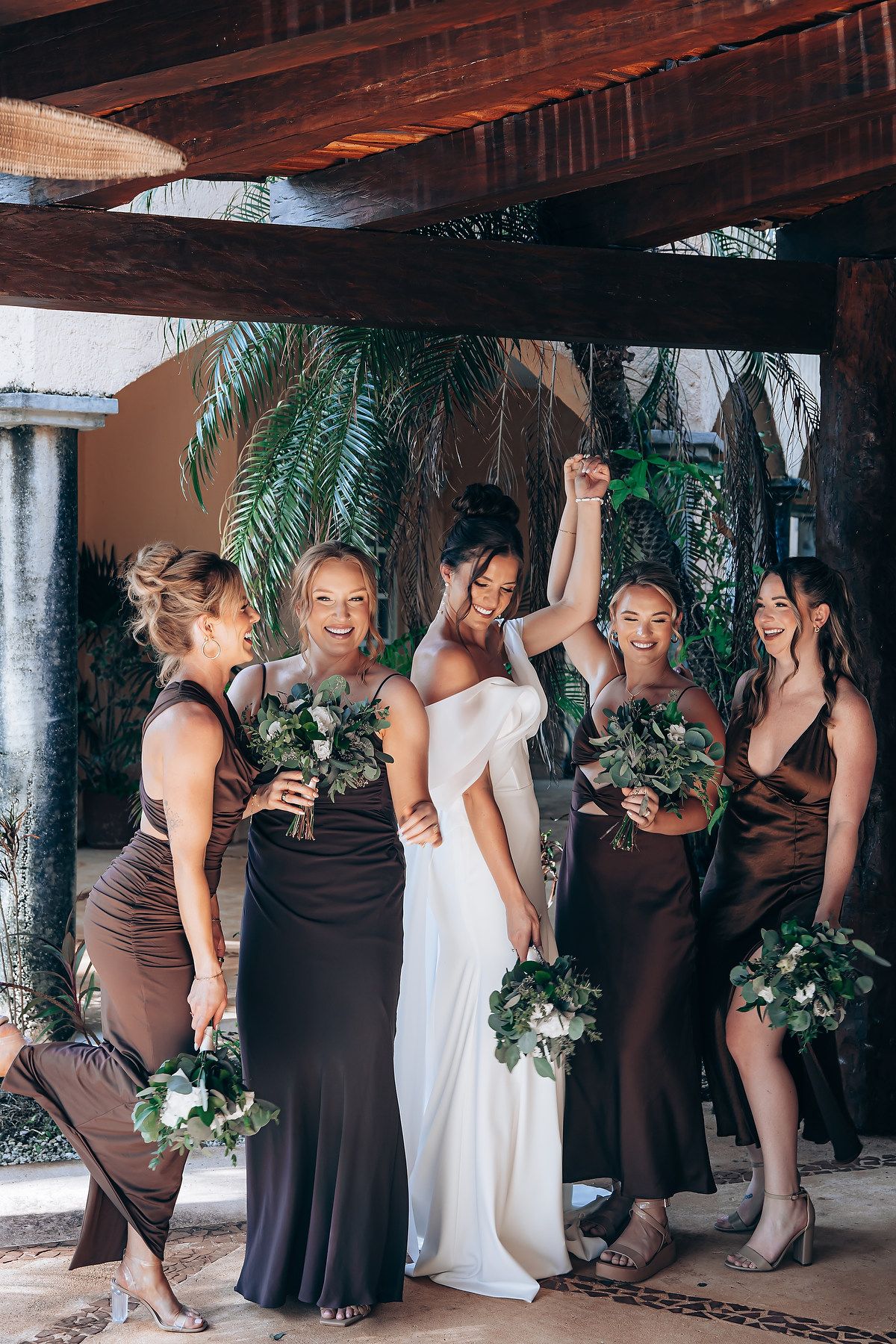 Bridesmaids in brown dresses pose together holding greenery bouquets under a wooden pergola.