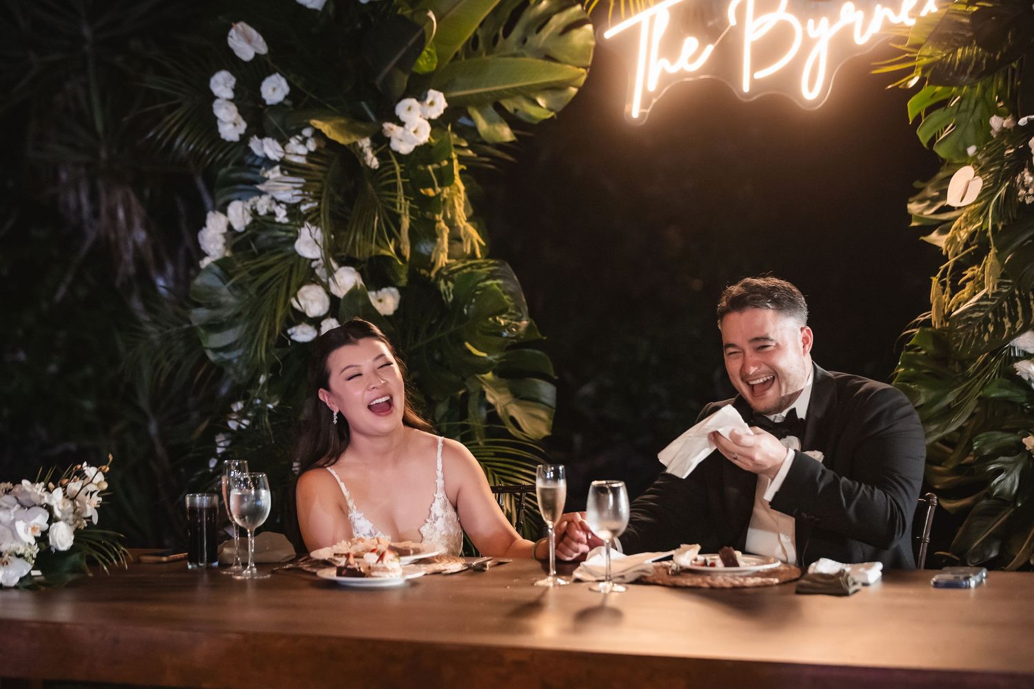 Wedding couple seated at illuminated reception table with tropical decor and neon signage.
