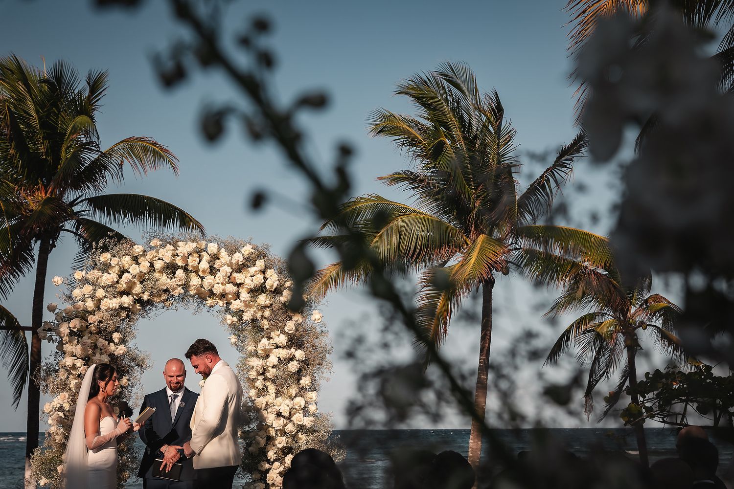 Romantic sunset wedding ceremony under white floral arch with palm trees silhouetted against sky.
