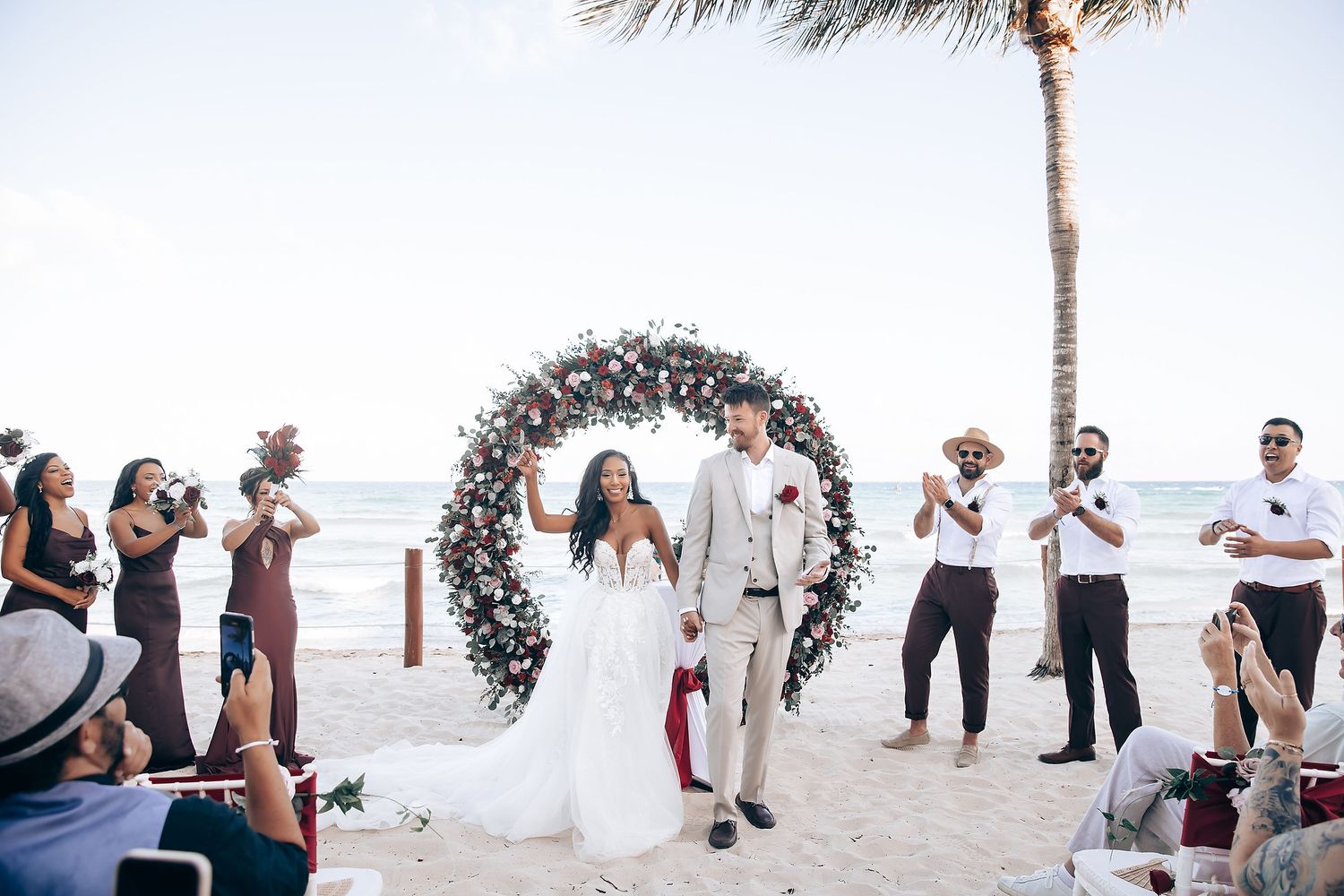 Beachfront wedding ceremony setup with white chairs and floral arrangements under palm trees.