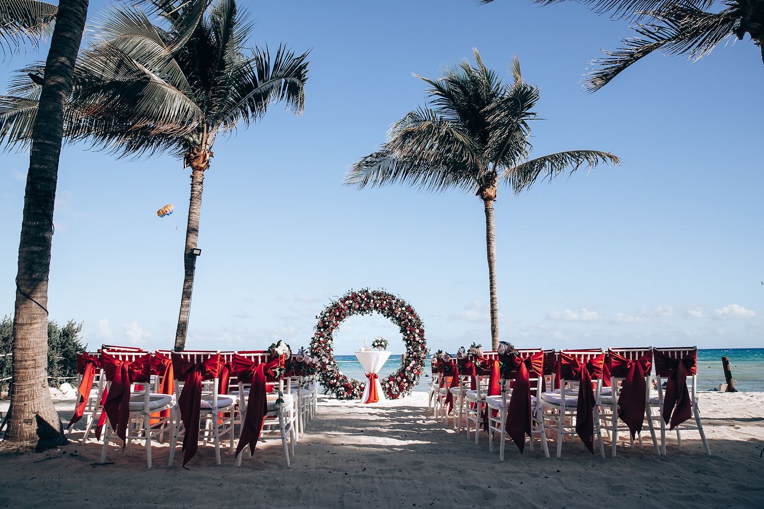 Panoramic beach wedding setup with palm trees and red chairs under clear blue skies.