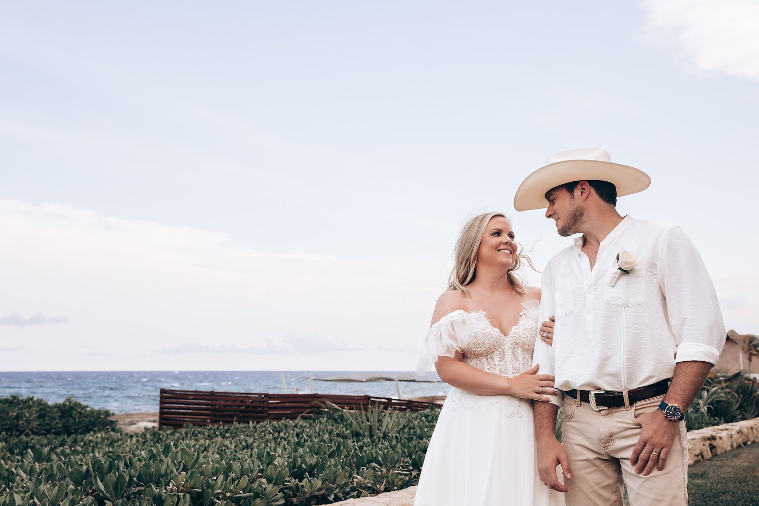Romantic couple in white outfits embrace while standing in a scenic outdoor landscape with mountains.