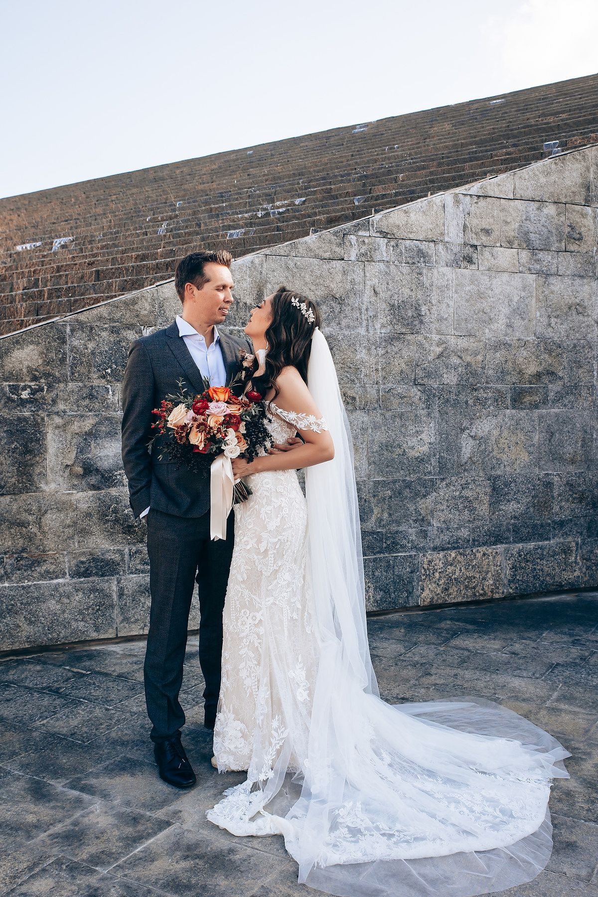 A wedding couple poses against a stone wall, with the bride wearing a lace gown with long train and veil.
