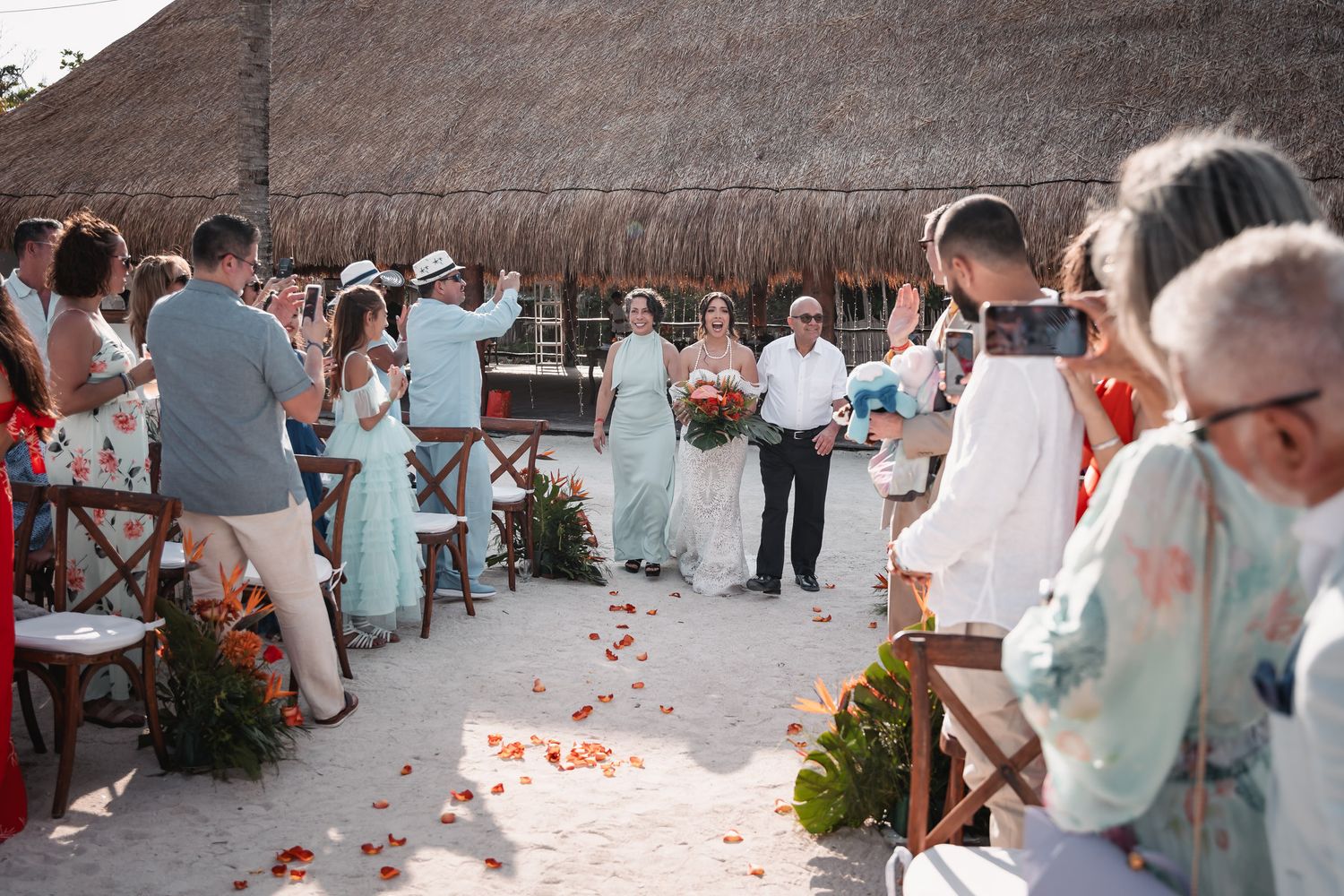 Bride walks down sandy aisle with escort as guests photograph beach wedding ceremony.