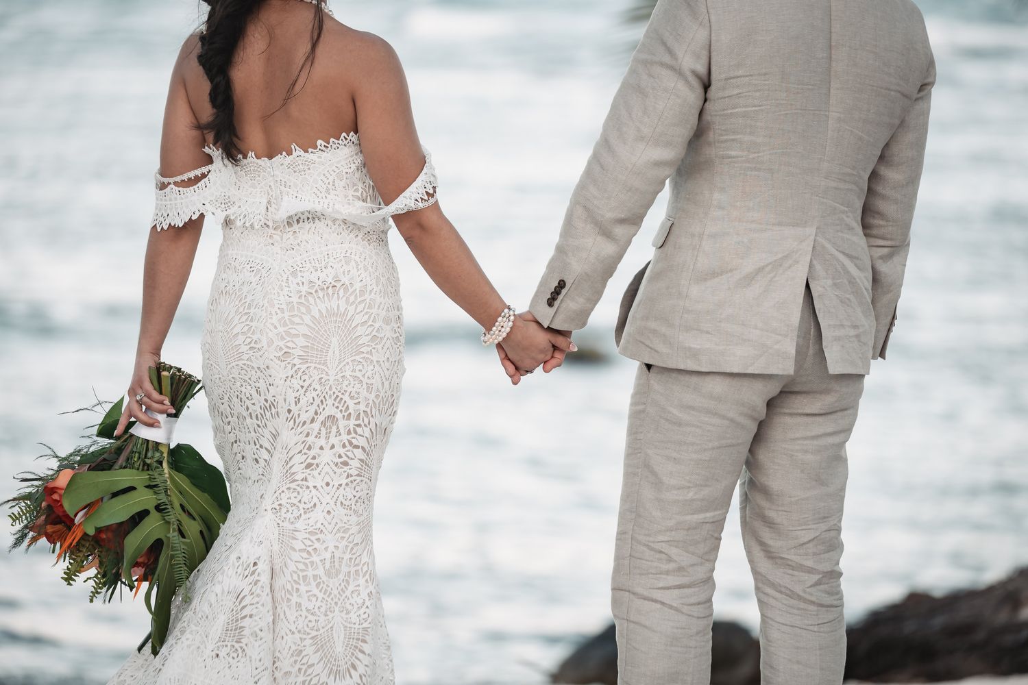 Bride holding tropical bouquet and groom holding hands facing ocean waves.