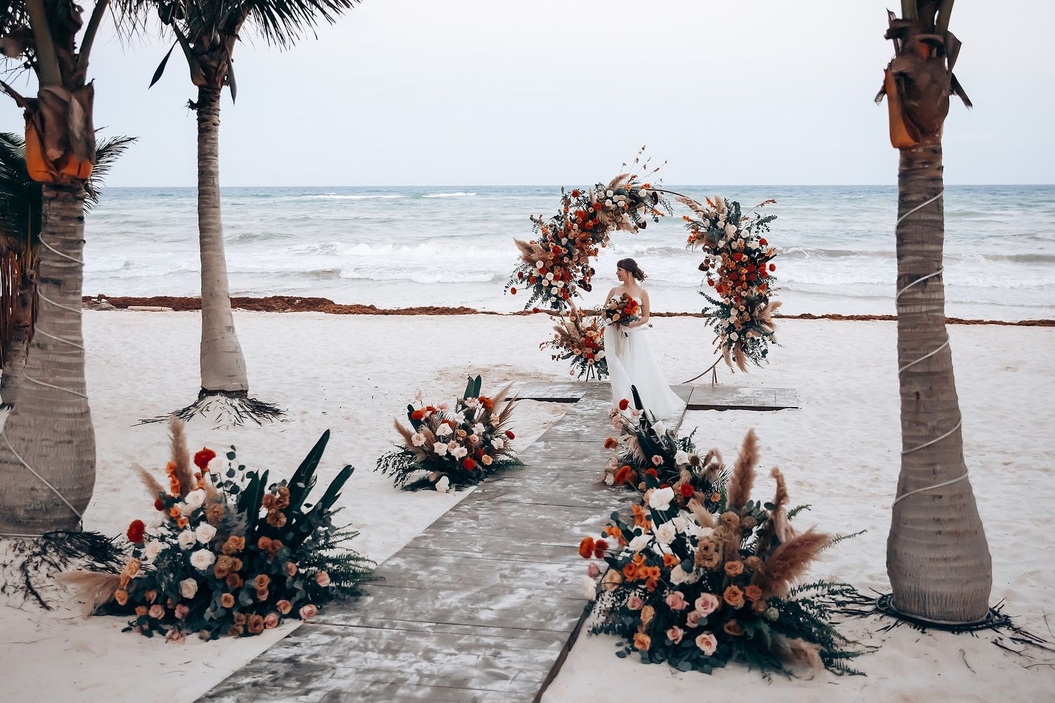 Tropical beach wedding ceremony setup with floral arrangements and palm trees along a sandy shoreline.