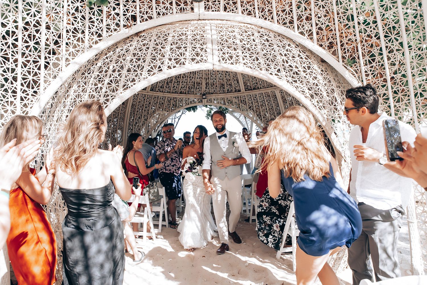 Wedding guests gathered under ornate latticed canopy during daytime celebration.