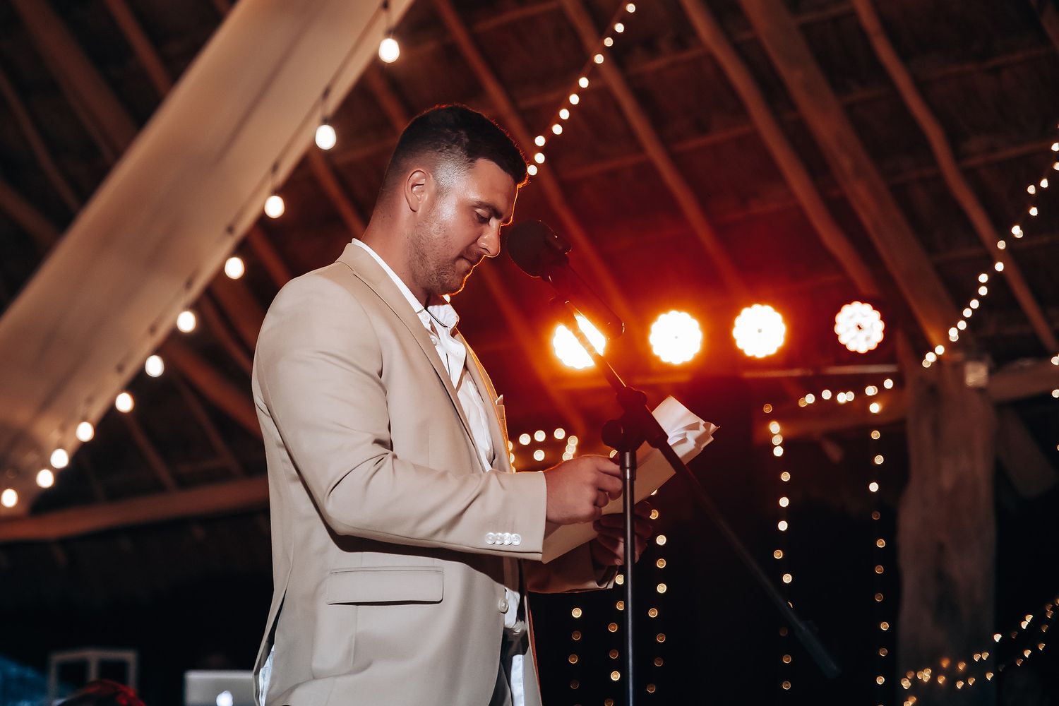 A person in a beige suit jacket speaks into a microphone against a backdrop of string lights and red stage lighting.