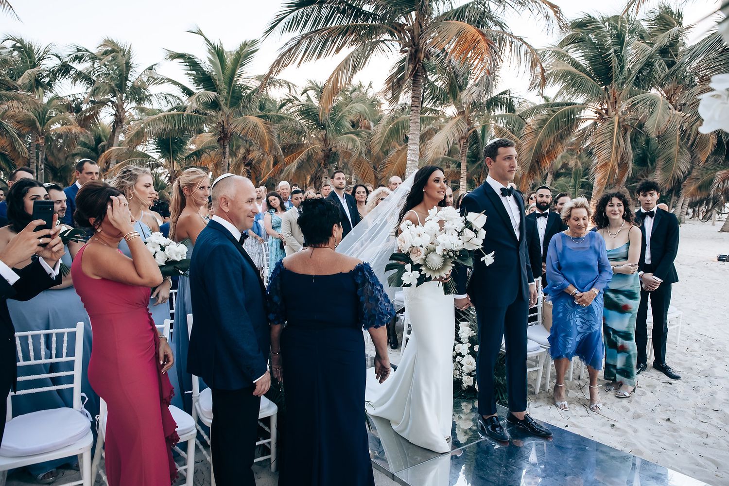 A beach wedding ceremony with guests gathered under palm trees.