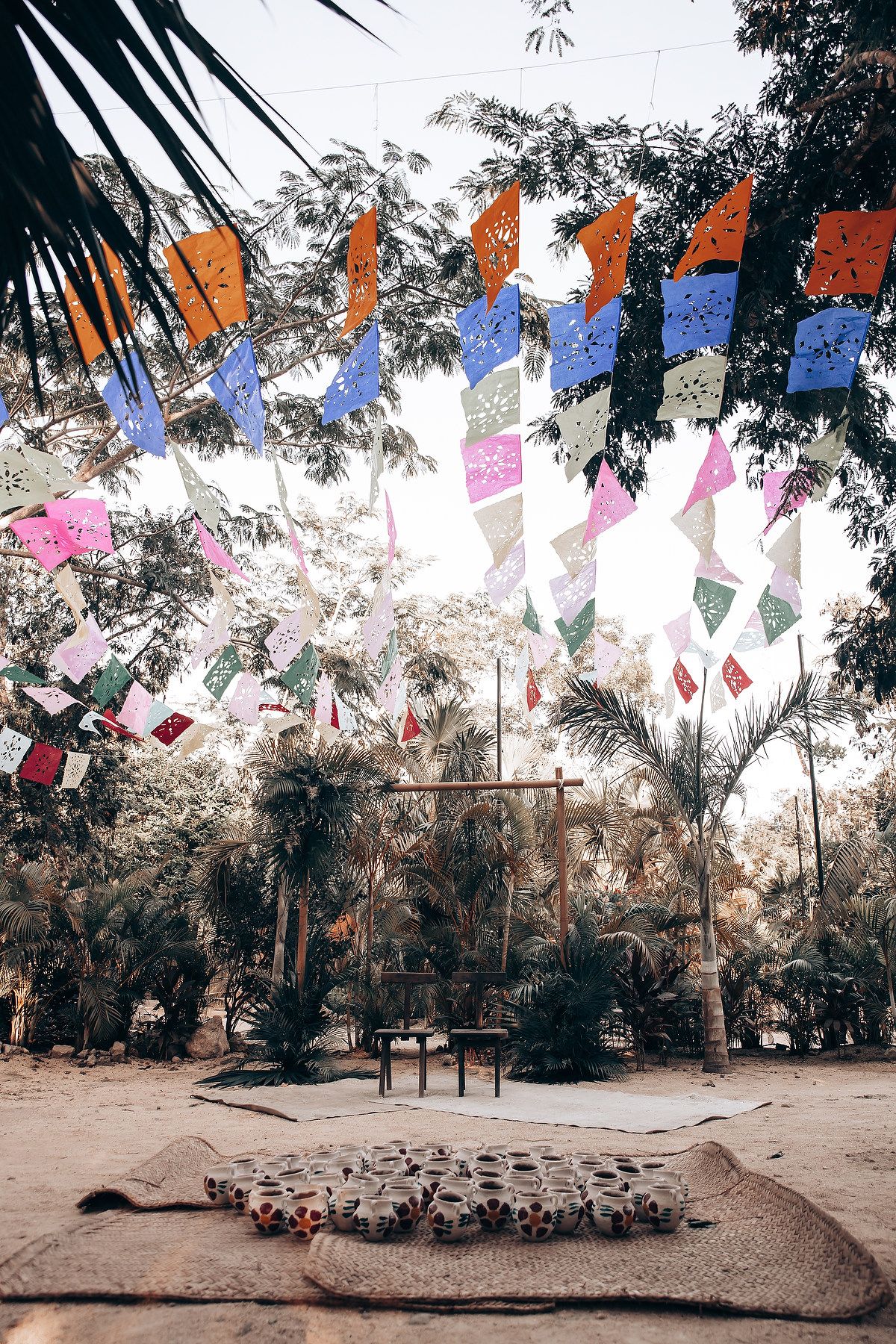 Colorful prayer flags hang between palm trees over a rustic outdoor ceremony space.