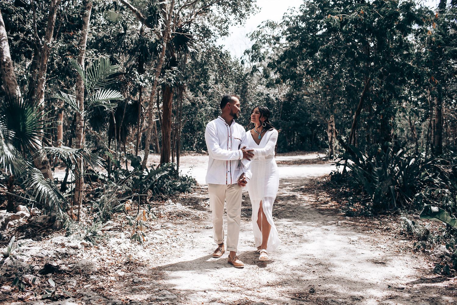 A couple in matching white outfits walks together along a sunlit forest path.