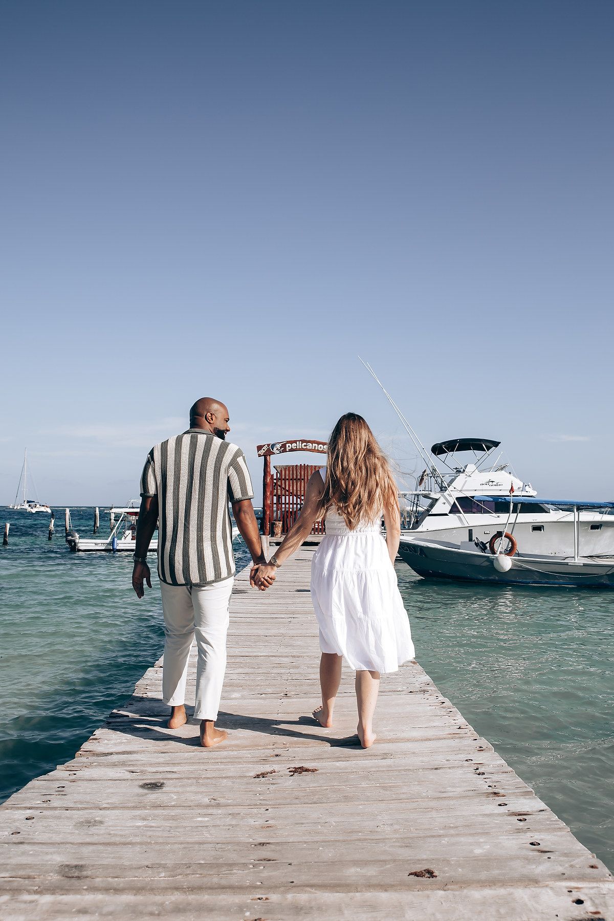 Interracial couple holding hands walking on a wooden pier with boats and turquoise ocean water.